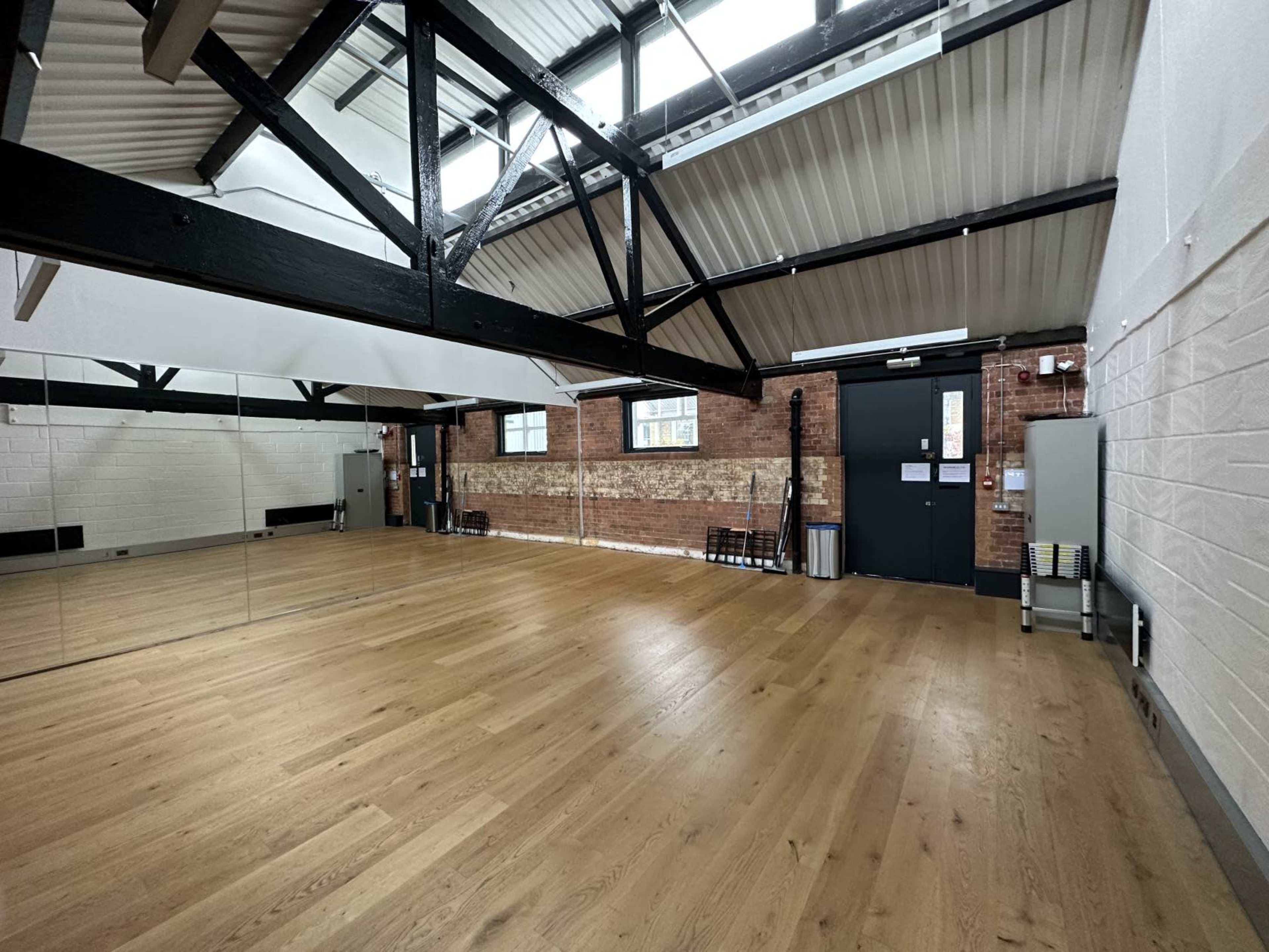 The image shows an empty room with wooden flooring, exposed brick walls, and large mirrors, featuring high ceilings supported by black beams.