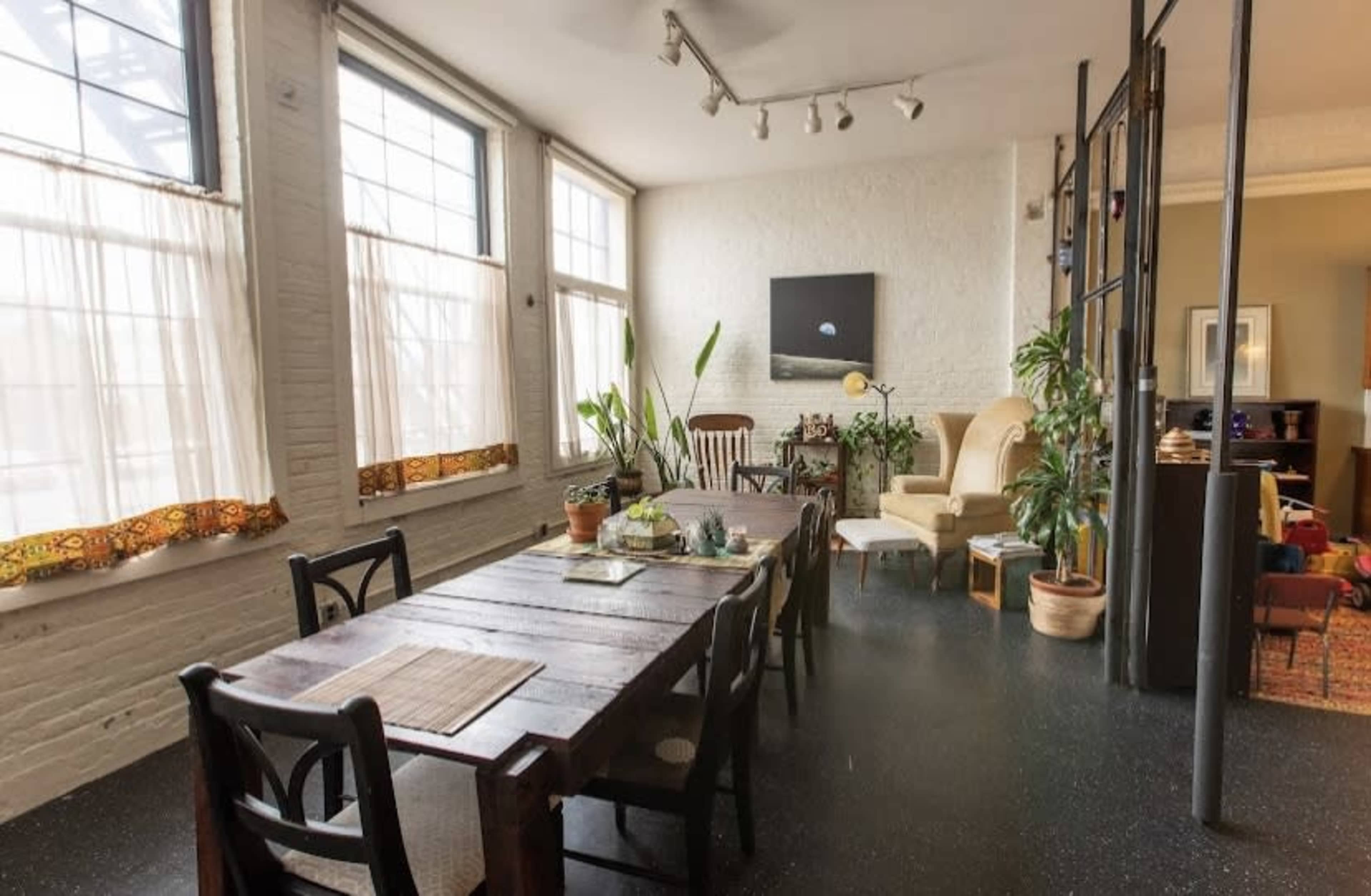 A spacious, bright living area features a wooden dining table, potted plants, and large windows with sheer curtains.
