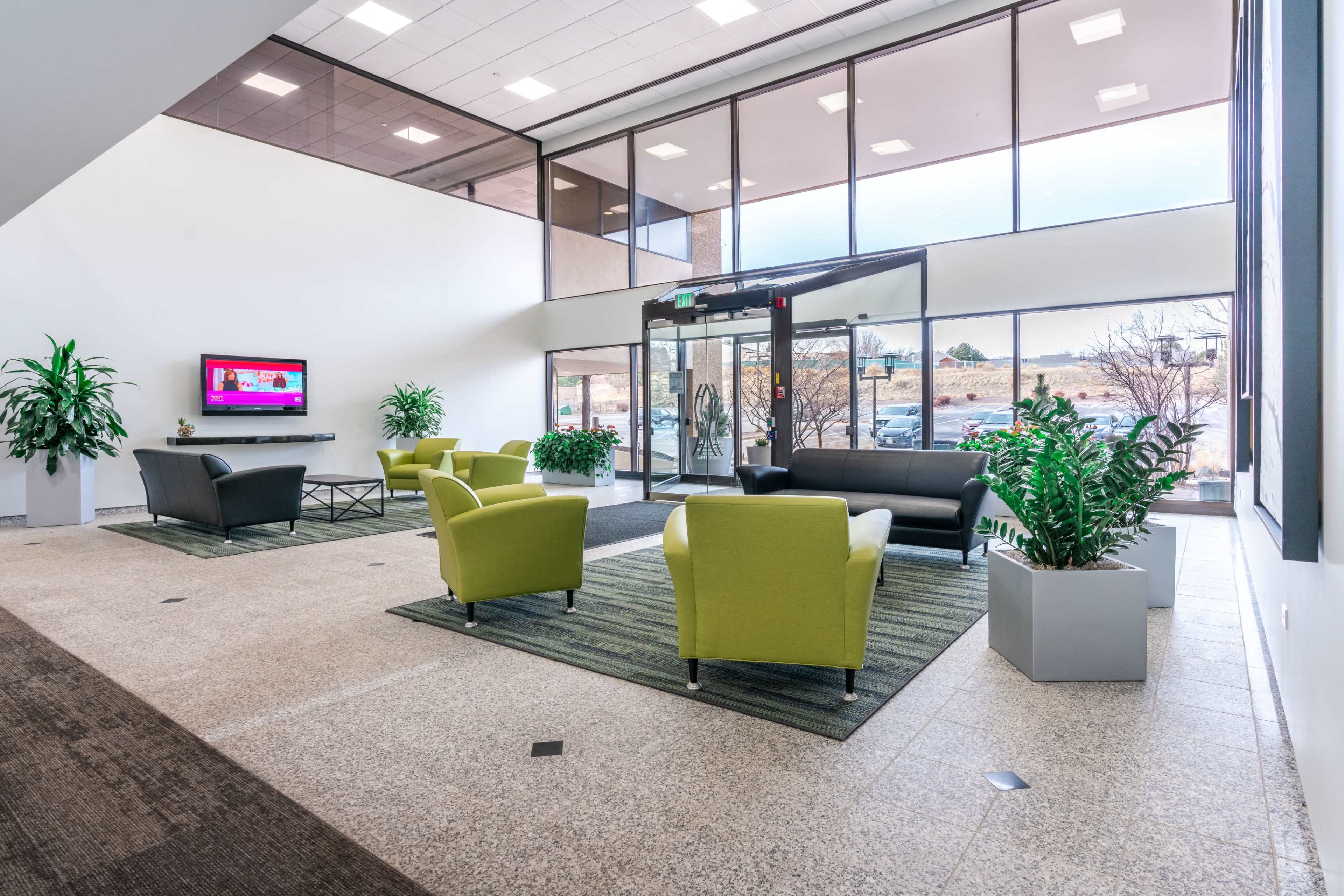 A modern lobby area with green chairs, a dark sofa, television, and large windows allowing natural light to enter.