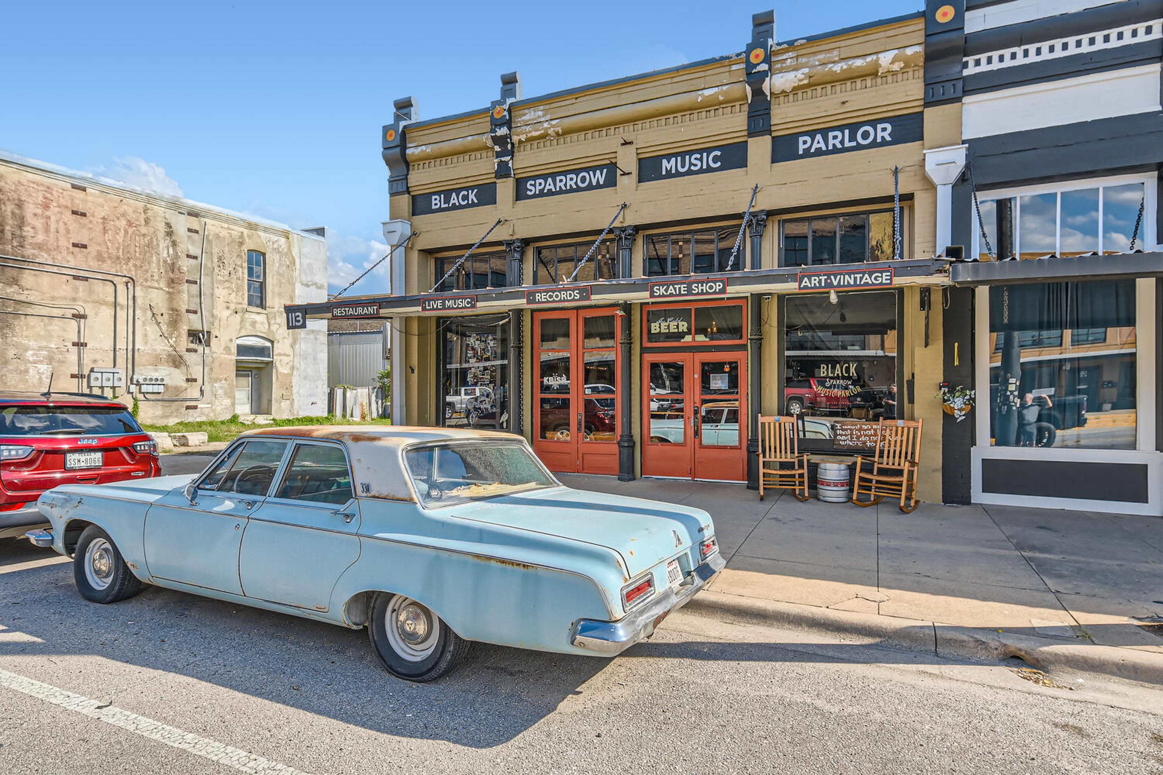 A light blue vintage car is parked in front of a music parlor labeled "Black Sparrow" on a sunny street with a mix of shops.