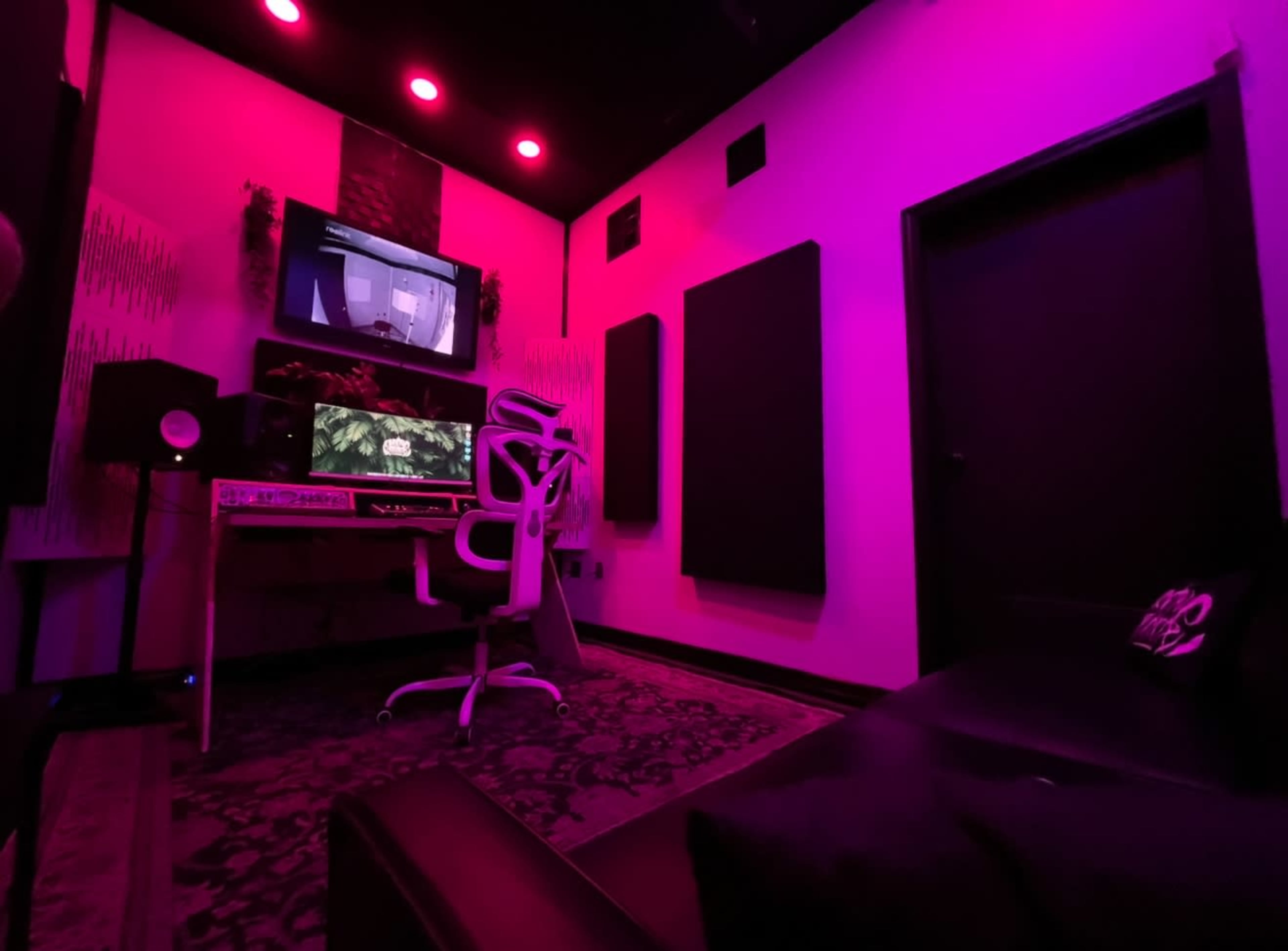 The image shows a dimly lit music studio featuring a desk with computer monitors, a chair, and soundproofing panels, all under pink lighting.