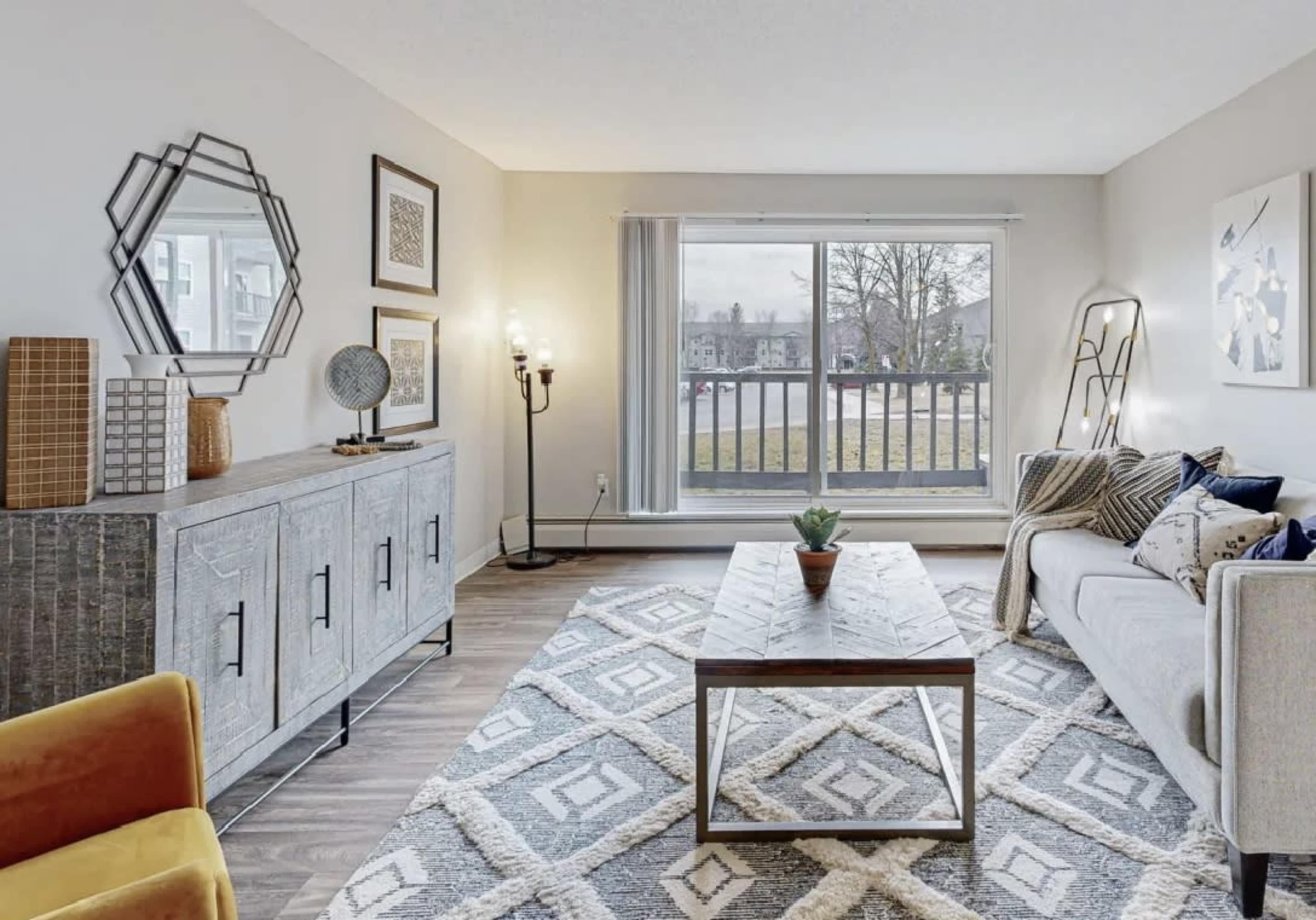A modern living room featuring a gray sofa, a coffee table, a patterned area rug, and a large window overlooking a grassy area.