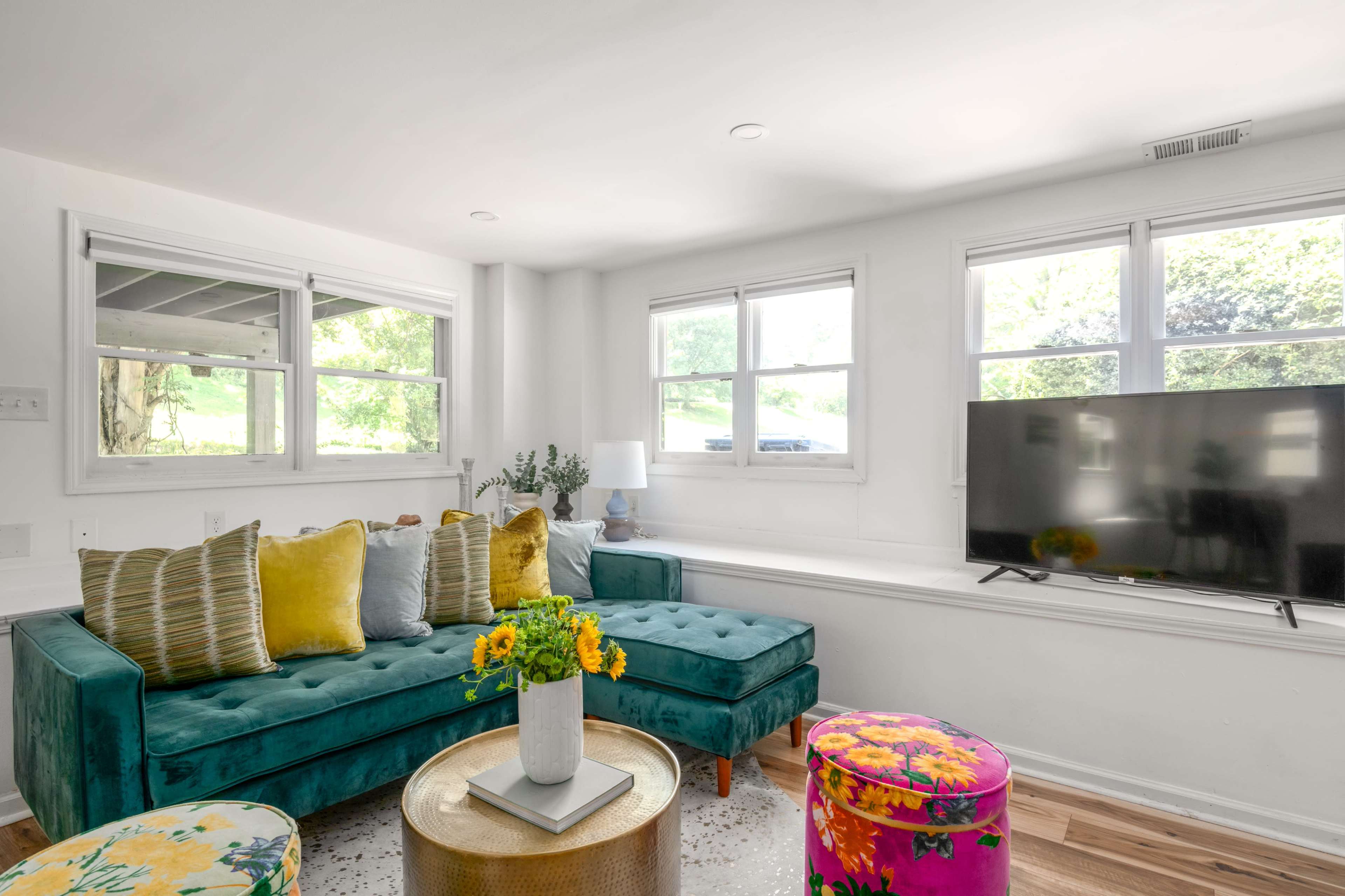 The image showcases a bright living room featuring a teal sofa with yellow and green pillows, a round coffee table, and a television against a white wall with large windows allowing natural light in.