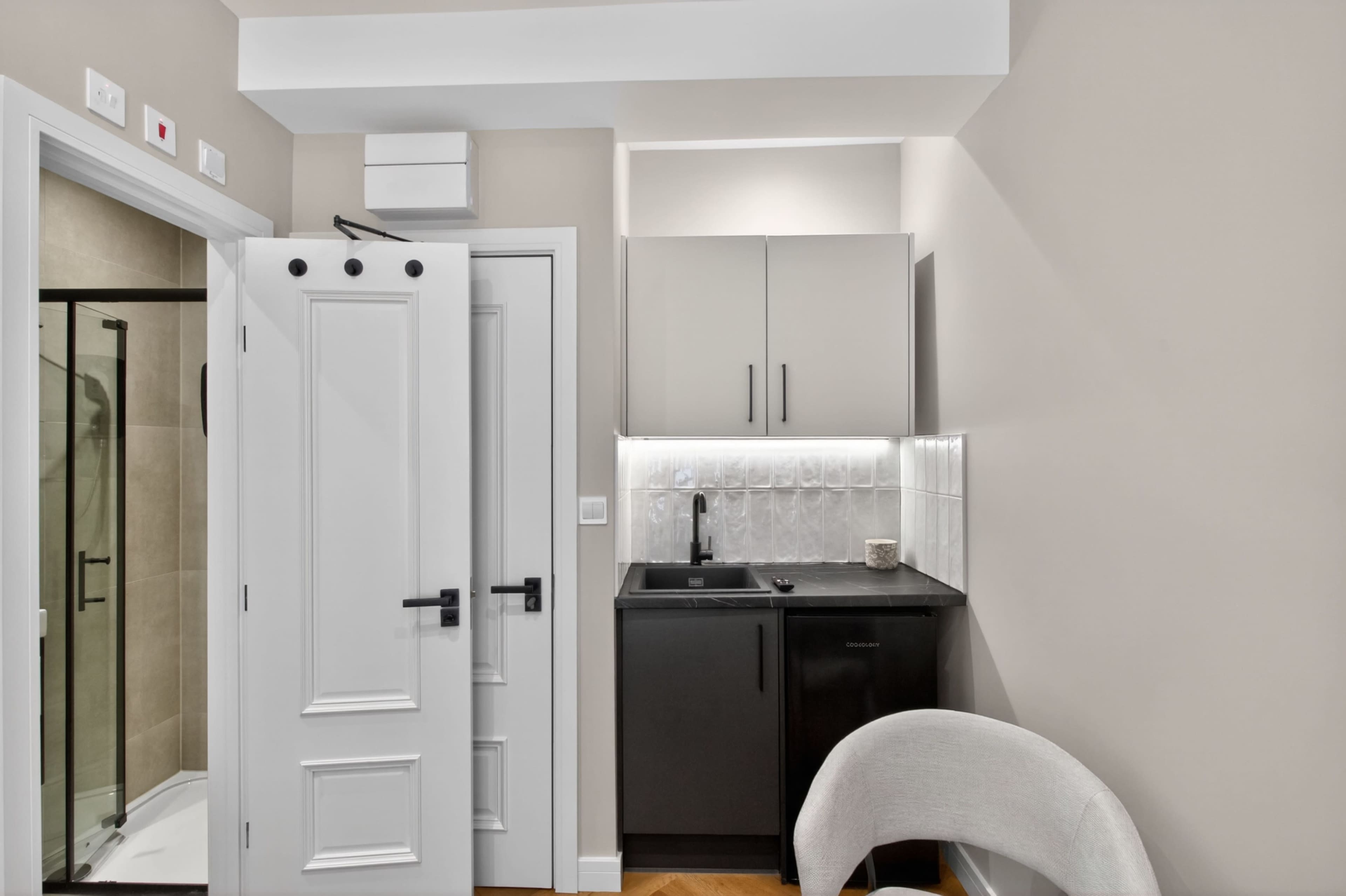 The image shows a modern kitchenette with a sink, black cabinets, and a white tiled backsplash, located next to a closed door in a light-colored room.
