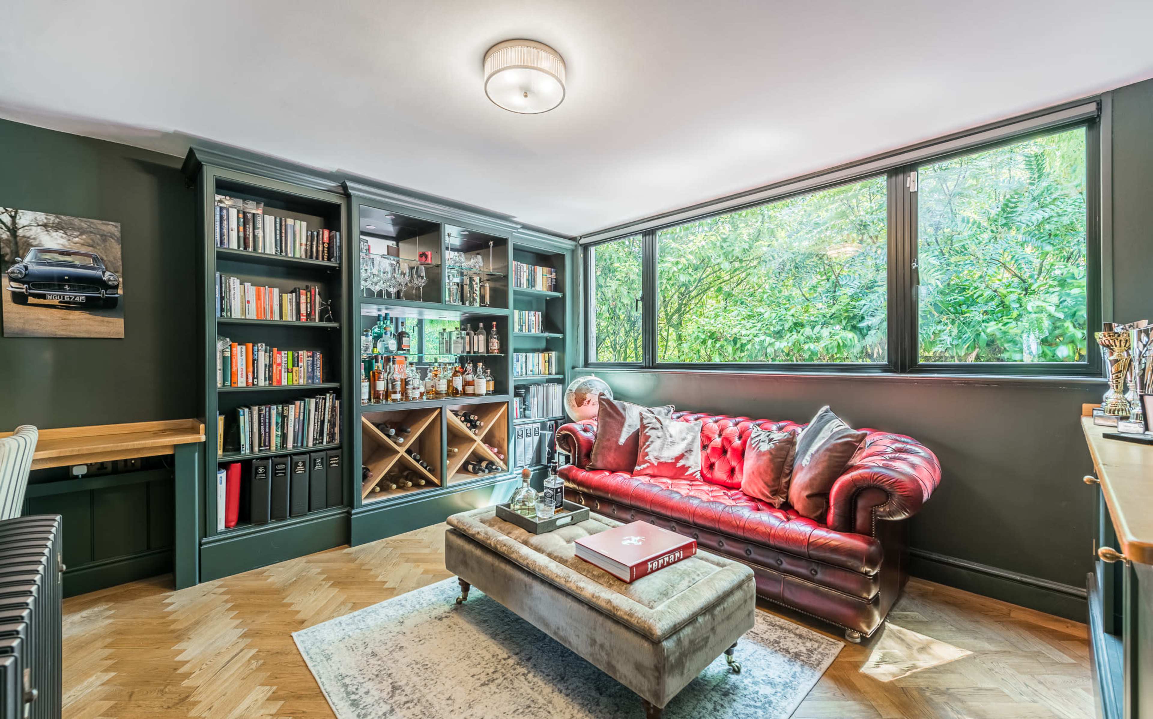 The image shows a cozy room featuring a red leather sofa, a coffee table, a bookshelf filled with various items, and large windows that overlook greenery.