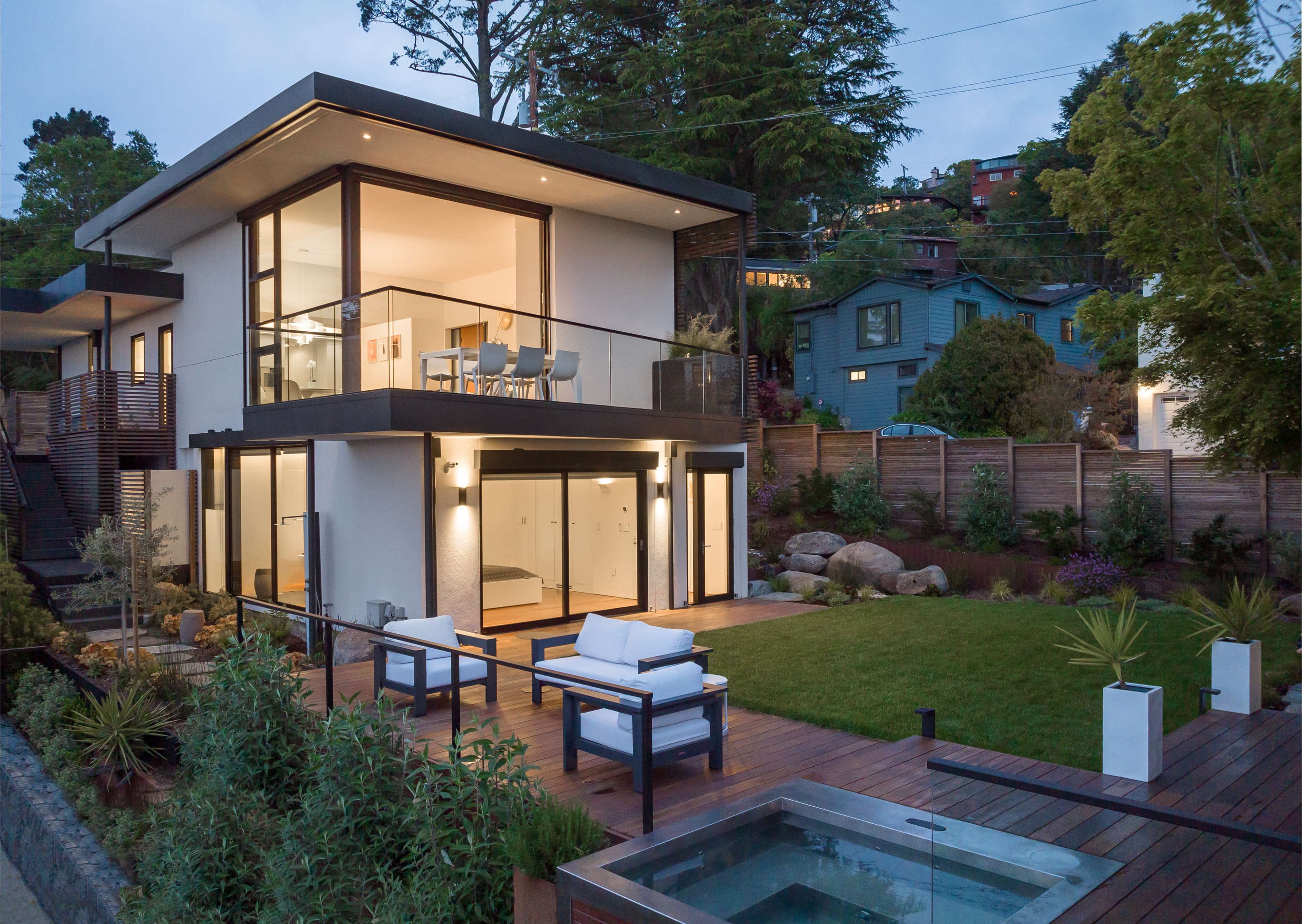 A modern two-story house features large glass windows, a patio with outdoor seating, and a landscaped yard with a hot tub.