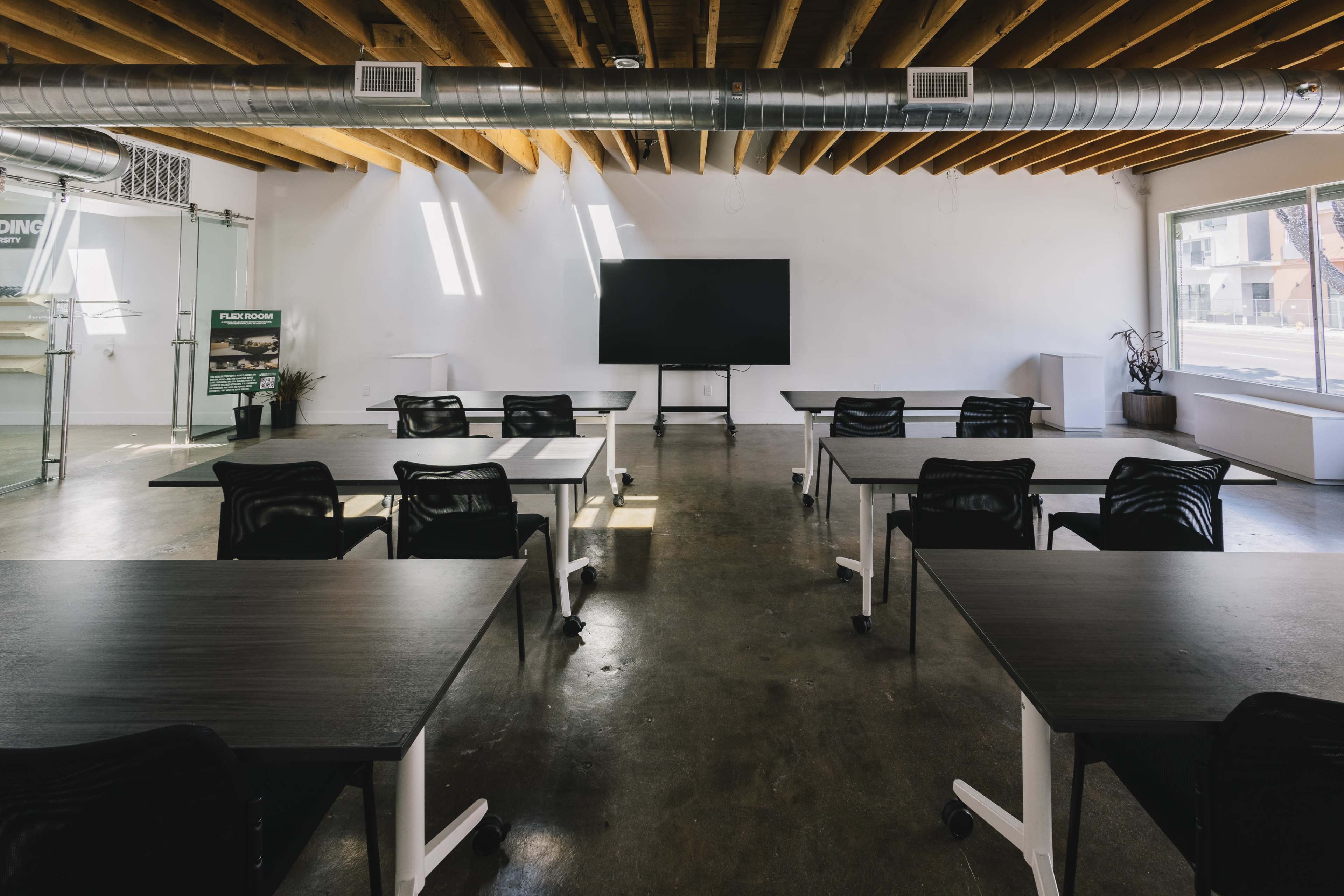 The image shows a modern, empty classroom with several black tables and chairs arranged in rows, featuring a large screen on one wall and windows allowing natural light to enter.