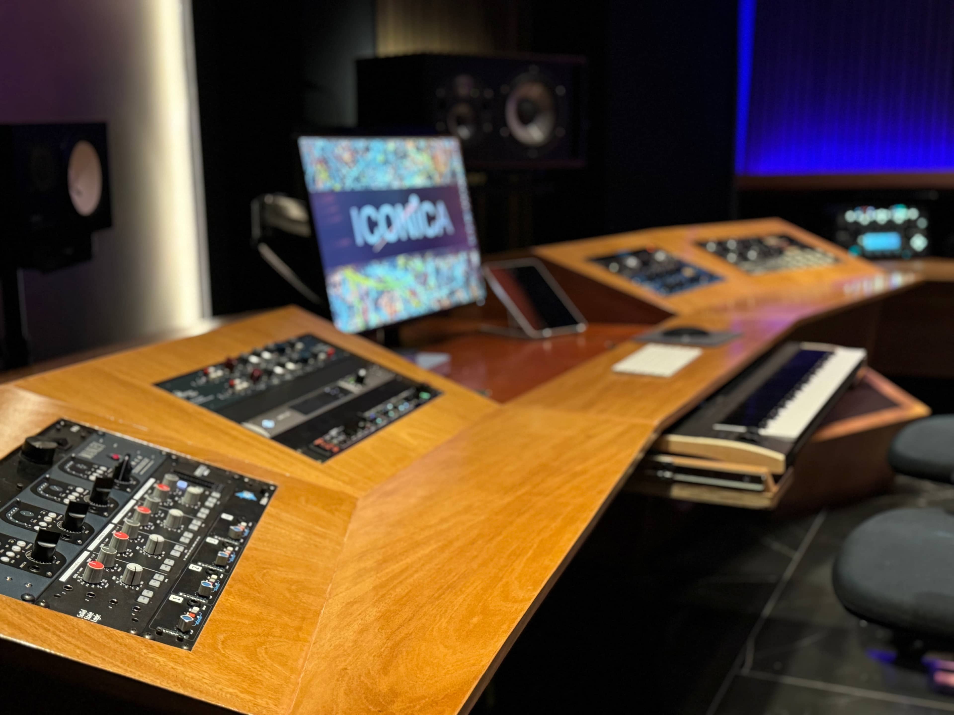 A modern music production studio with a wooden control desk, various audio equipment, a keyboard, and a computer monitor displaying colorful graphics.