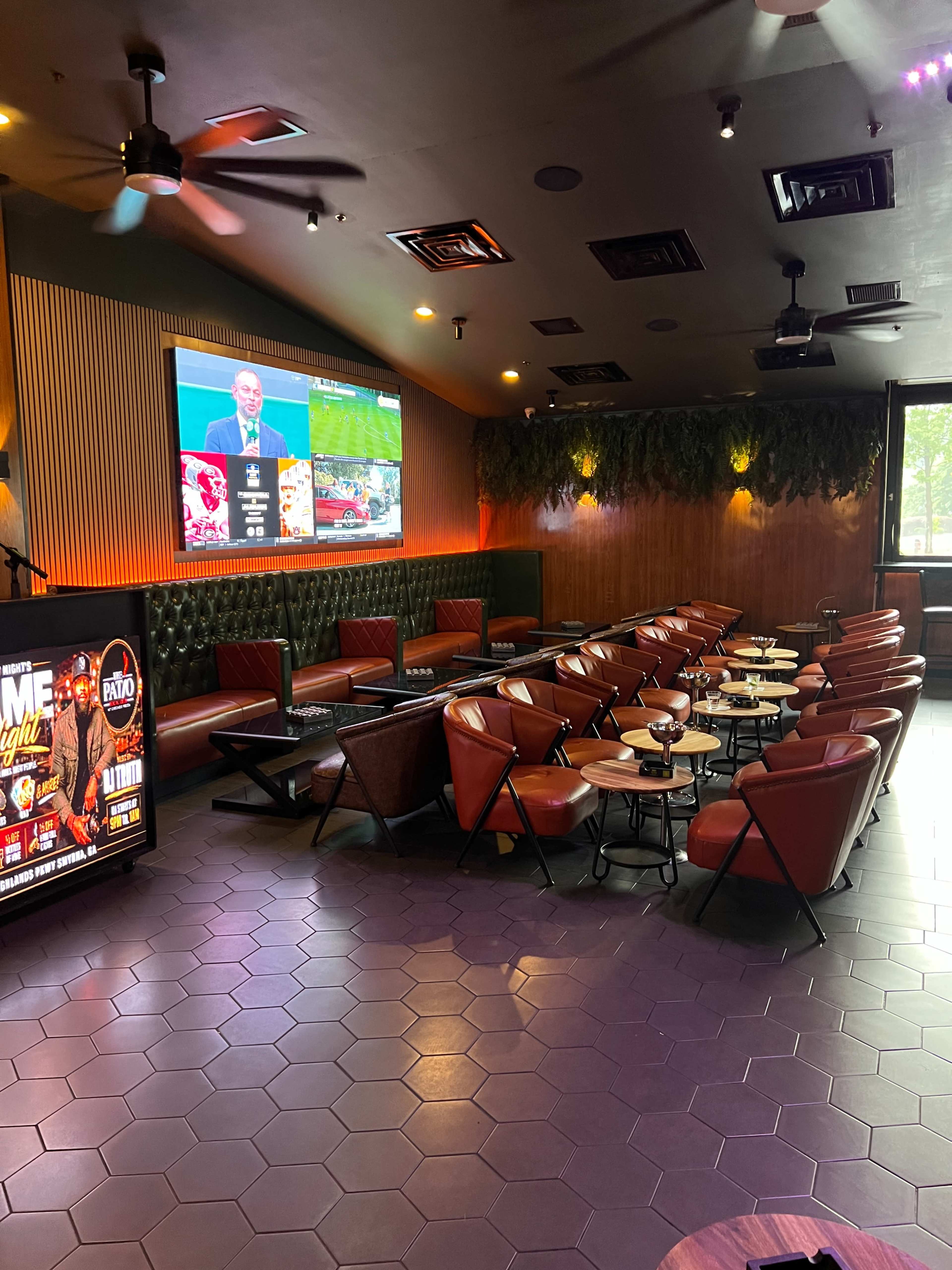 The interior of a sports lounge features multiple seating areas with red chairs and dark wooden tables, illuminated by a large screen displaying a sports game.