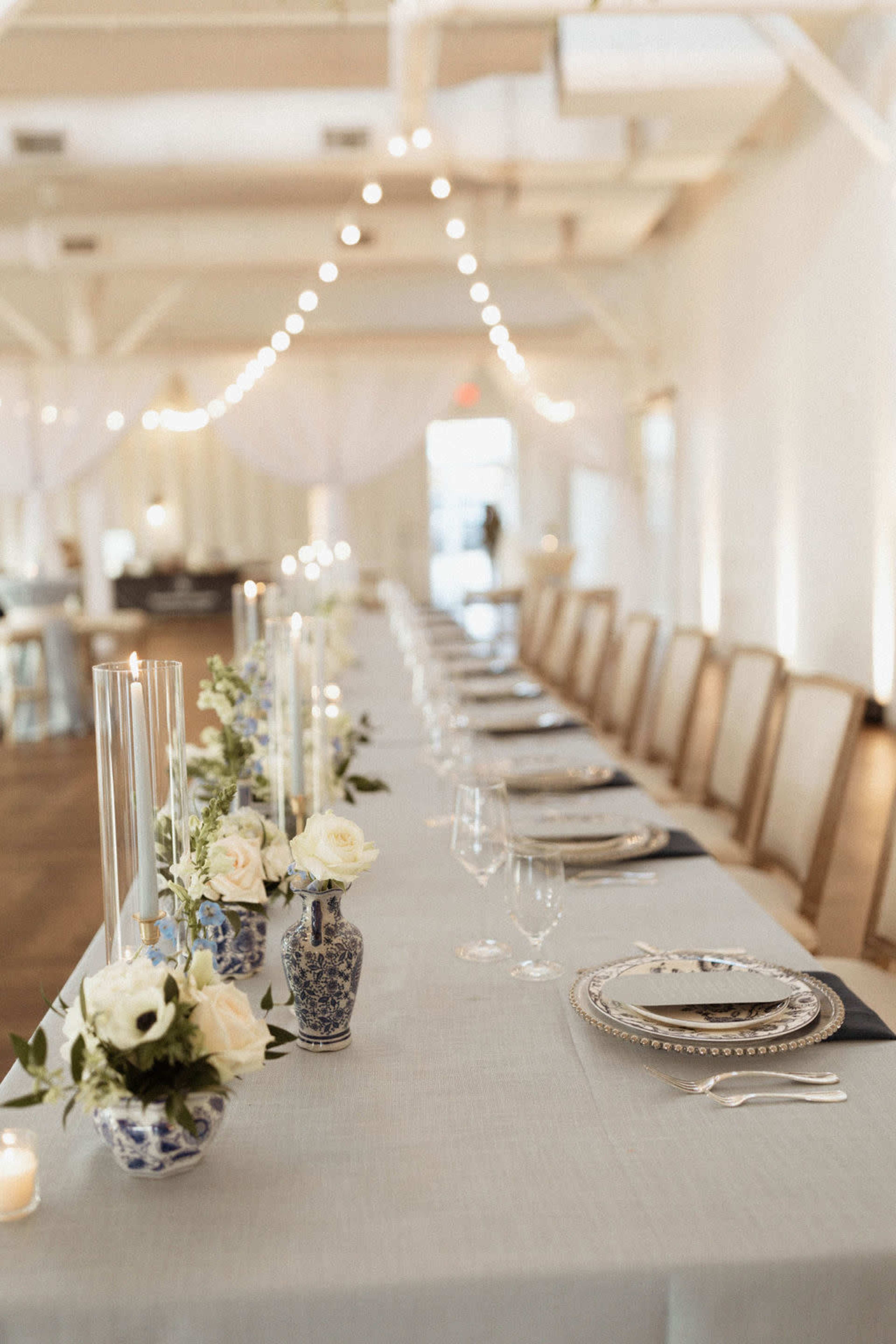 A long, elegantly set dining table is adorned with blue and white vases, candles, and floral arrangements under string lights in a bright, open space.