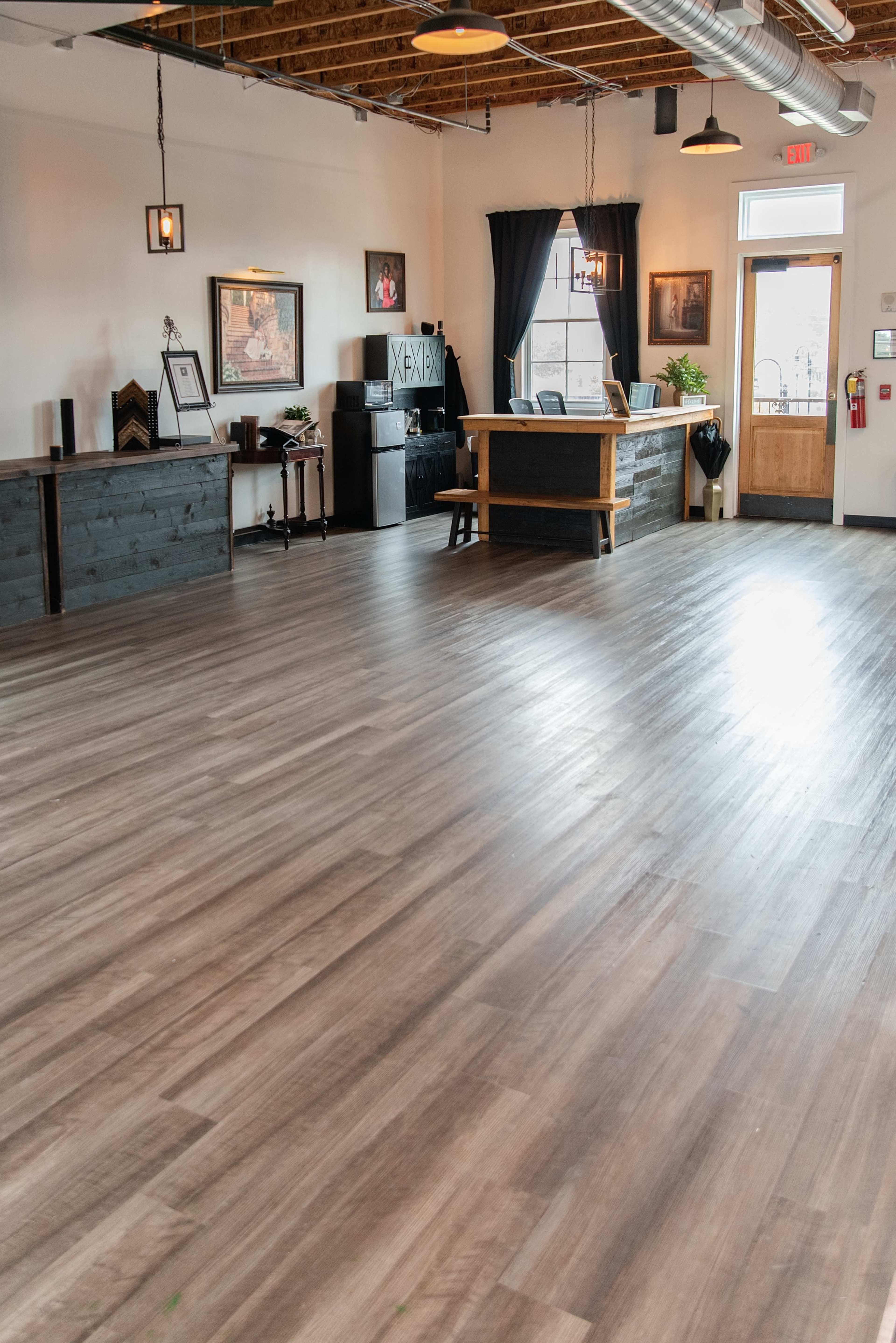 The image shows a spacious room with wooden flooring, featuring a reception area with a counter, several tables, and large windows letting in natural light.