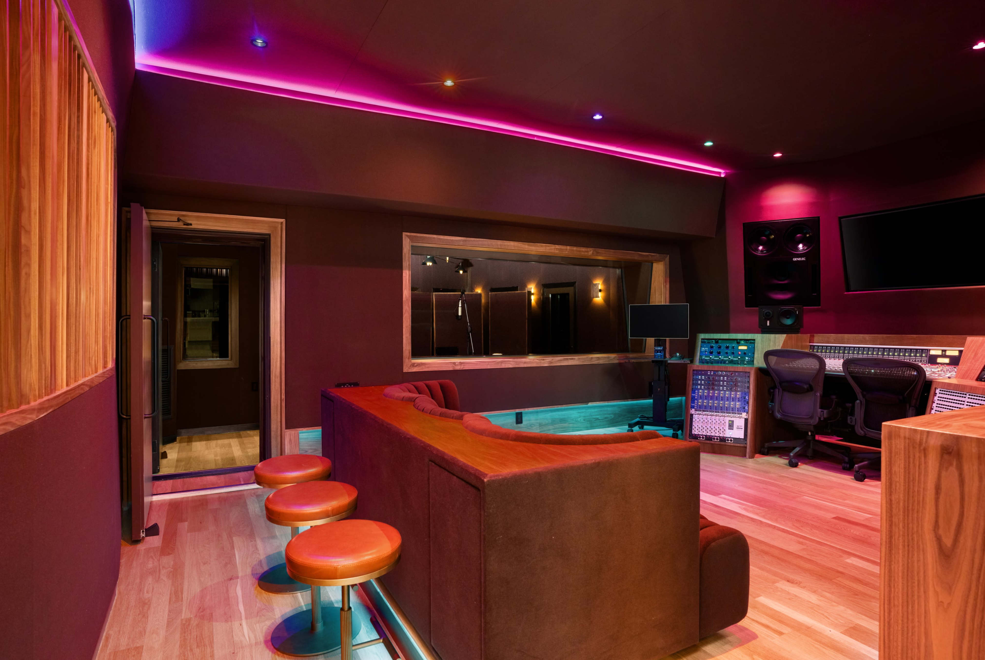 The image shows a modern recording studio featuring a sound mixing area with a large control panel, a viewing window, and a seating area with brown couches and orange bar stools.