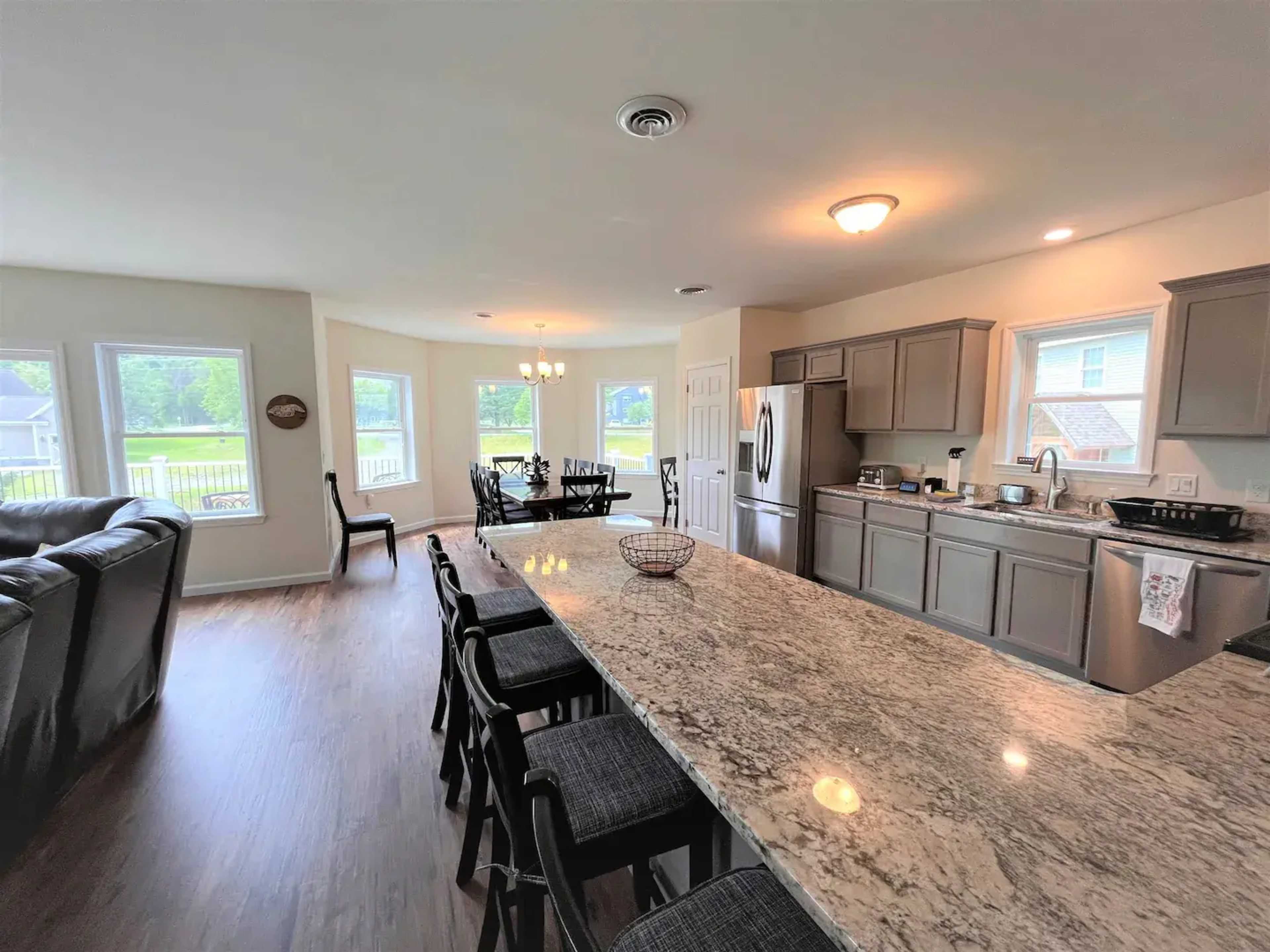 A spacious kitchen with gray cabinets, a large granite island with bar seating, and windows overlooking a green outdoor area.
