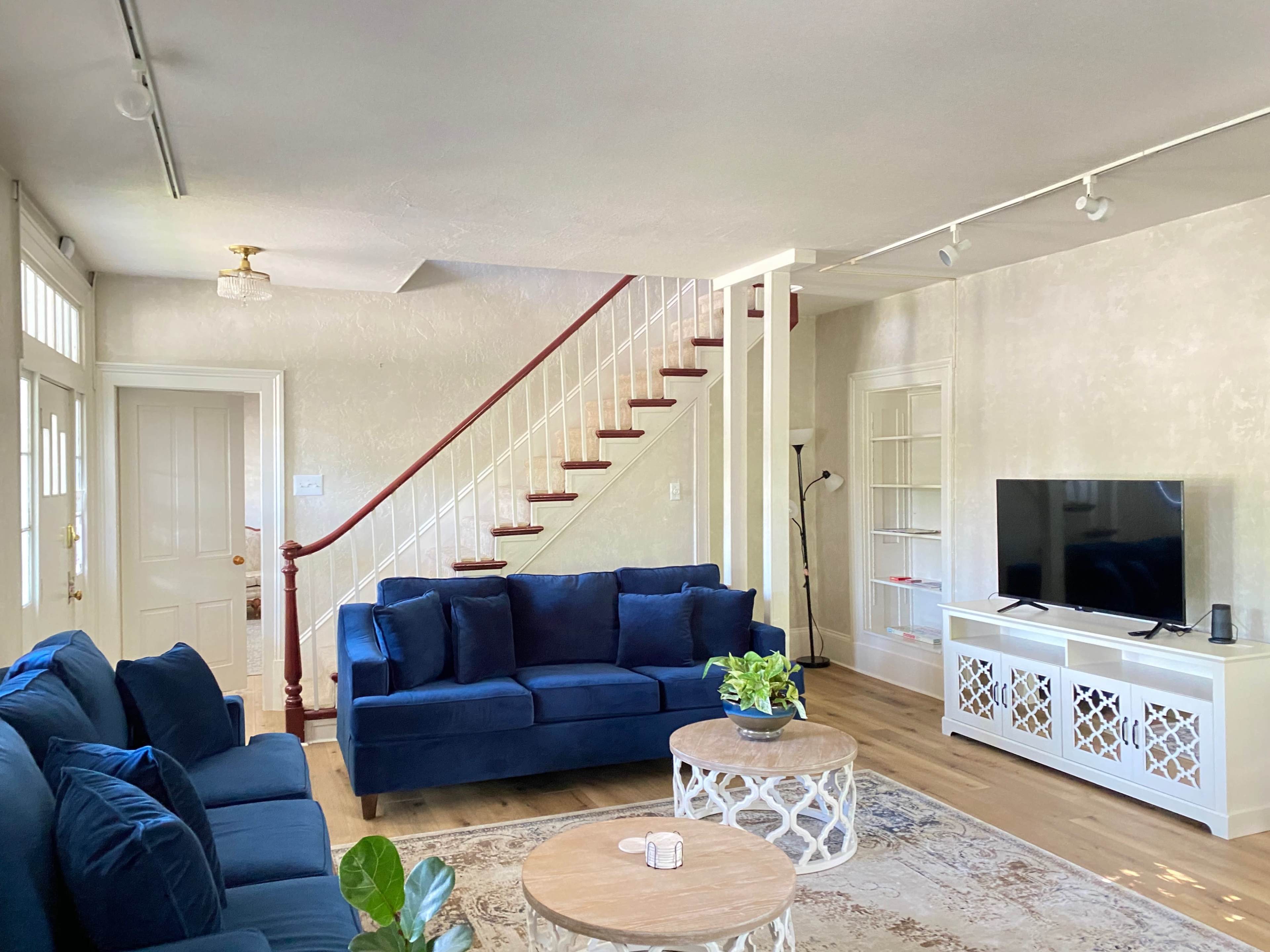 A living room with a blue sofa set, a round coffee table, a television on a white cabinet, and a staircase in the background.