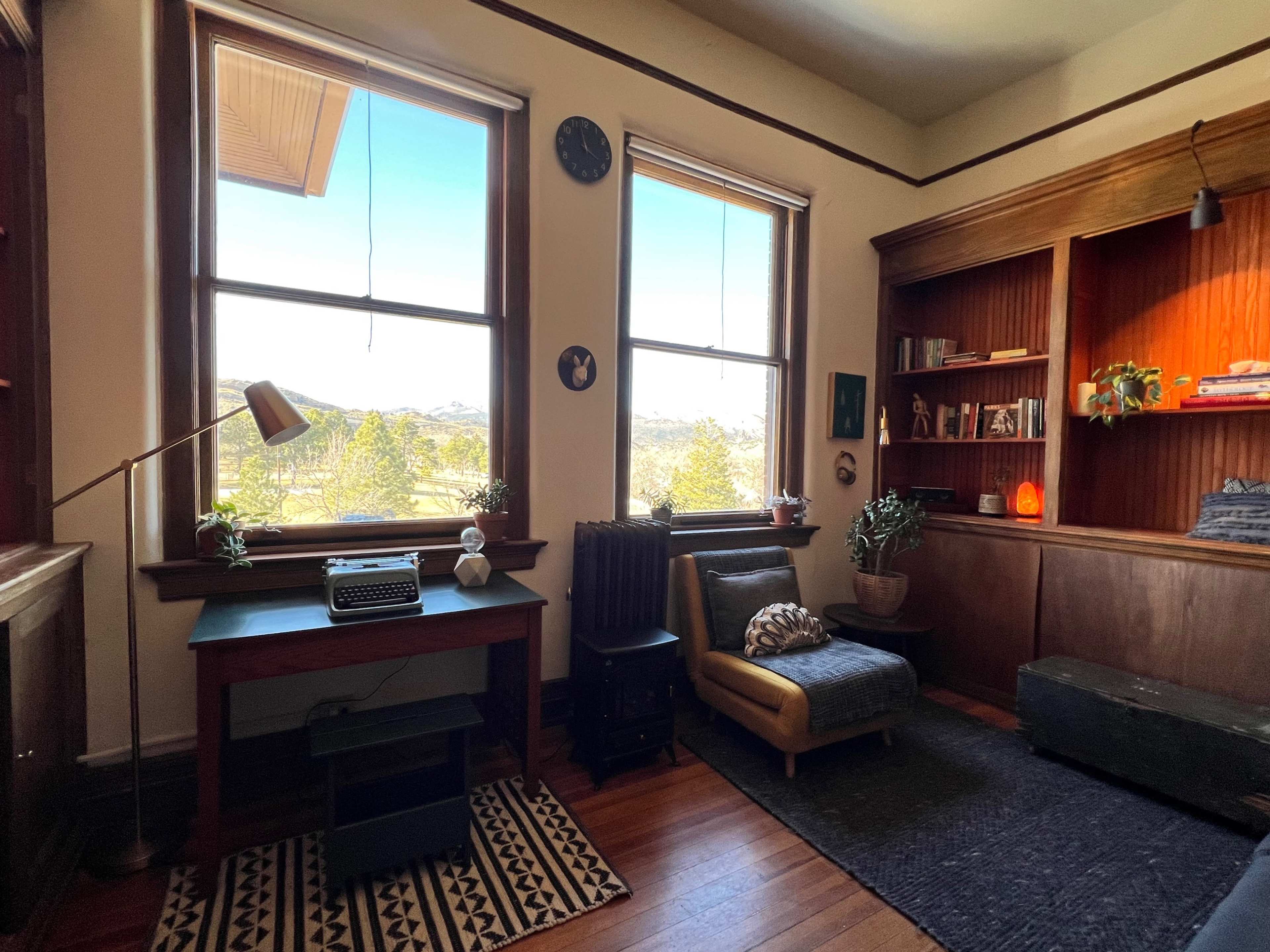 A cozy room with a wooden desk featuring a typewriter, a chair, bookshelves, and large windows offering a view of the outdoors.