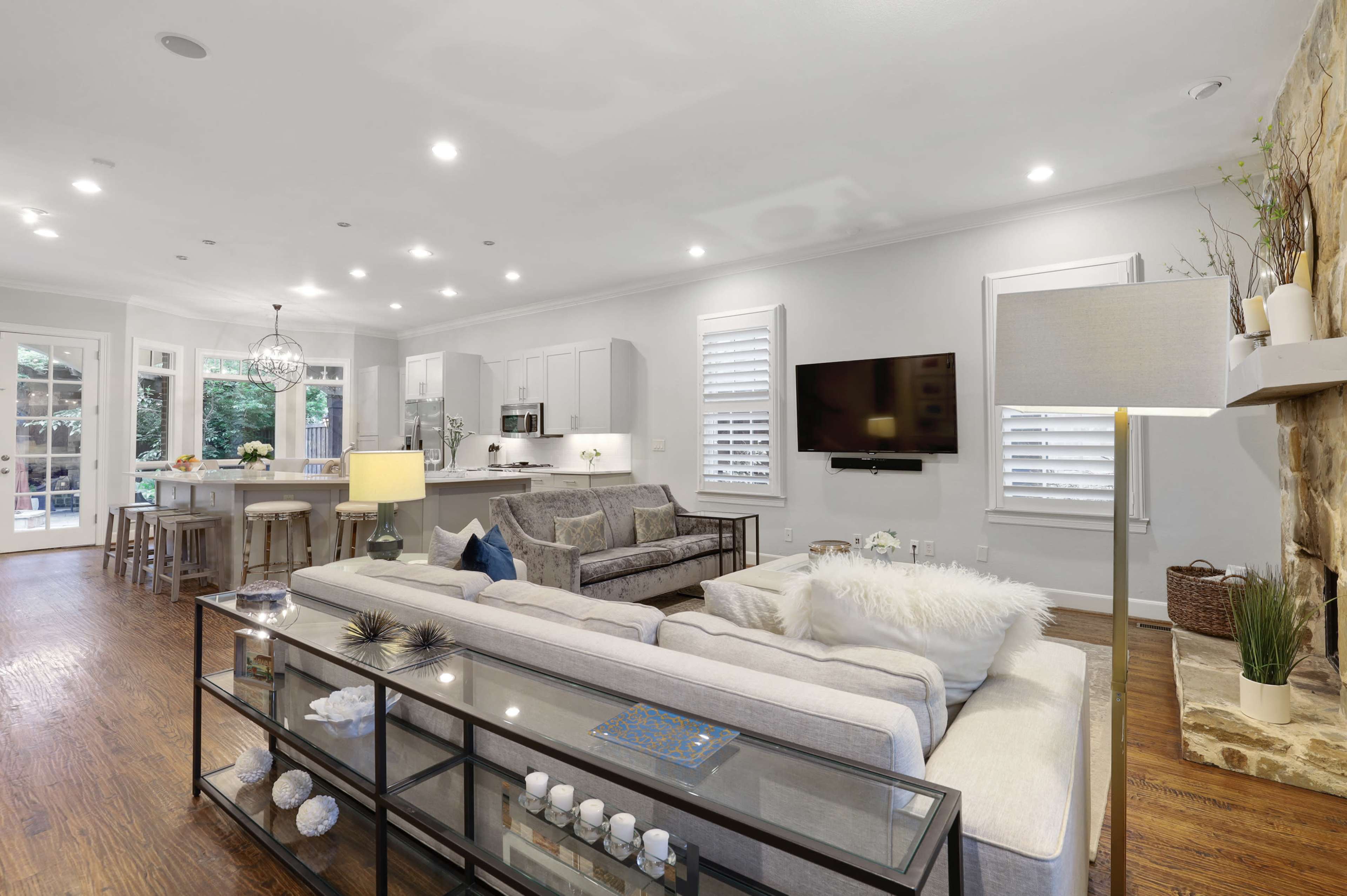 A modern living room features a light-colored sofa, a glass coffee table, and a view of a kitchen area with white cabinetry and a dining space.