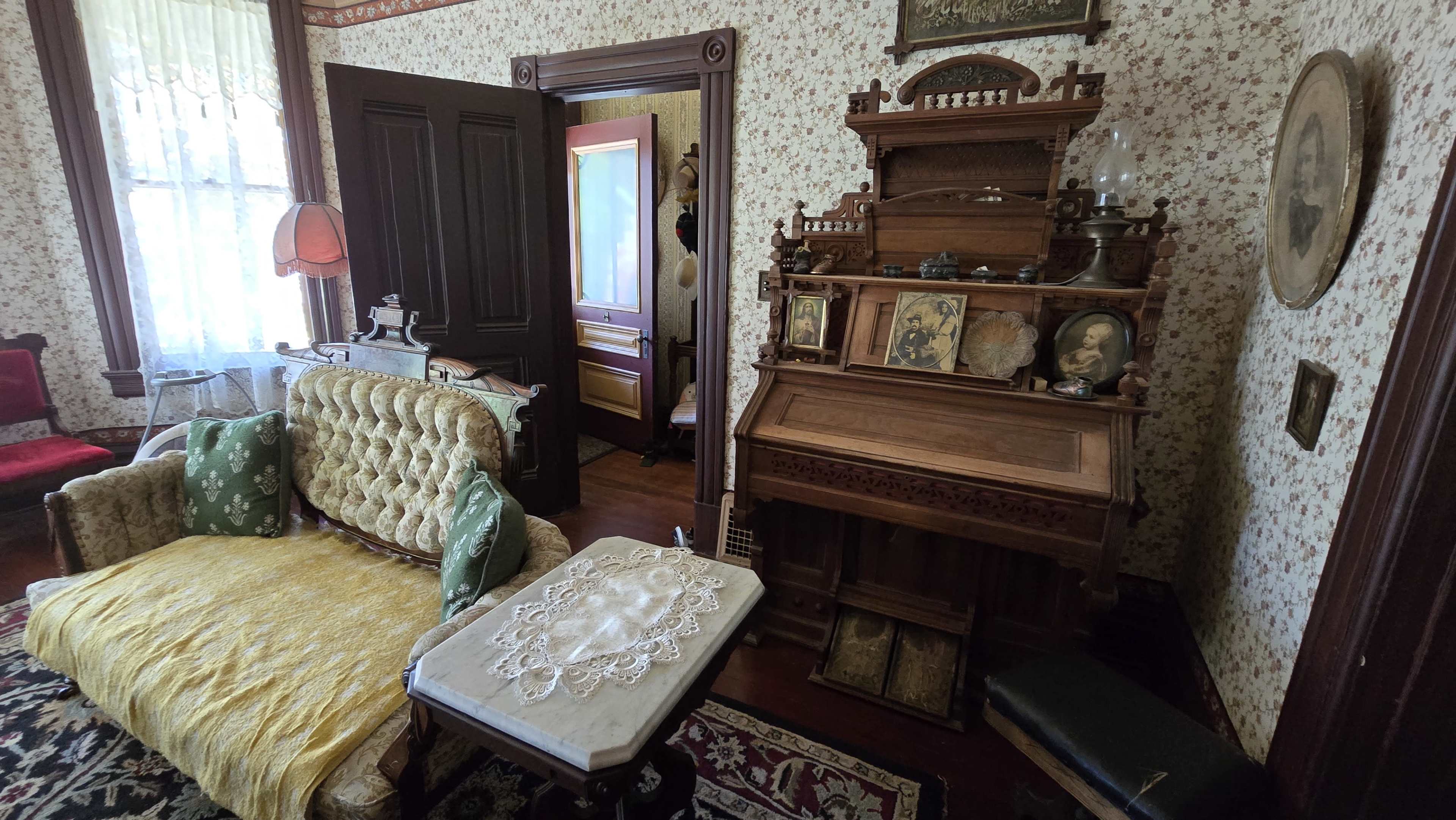 A vintage room featuring an ornate sofa, a wooden writing desk with framed photographs, and floral wallpaper.