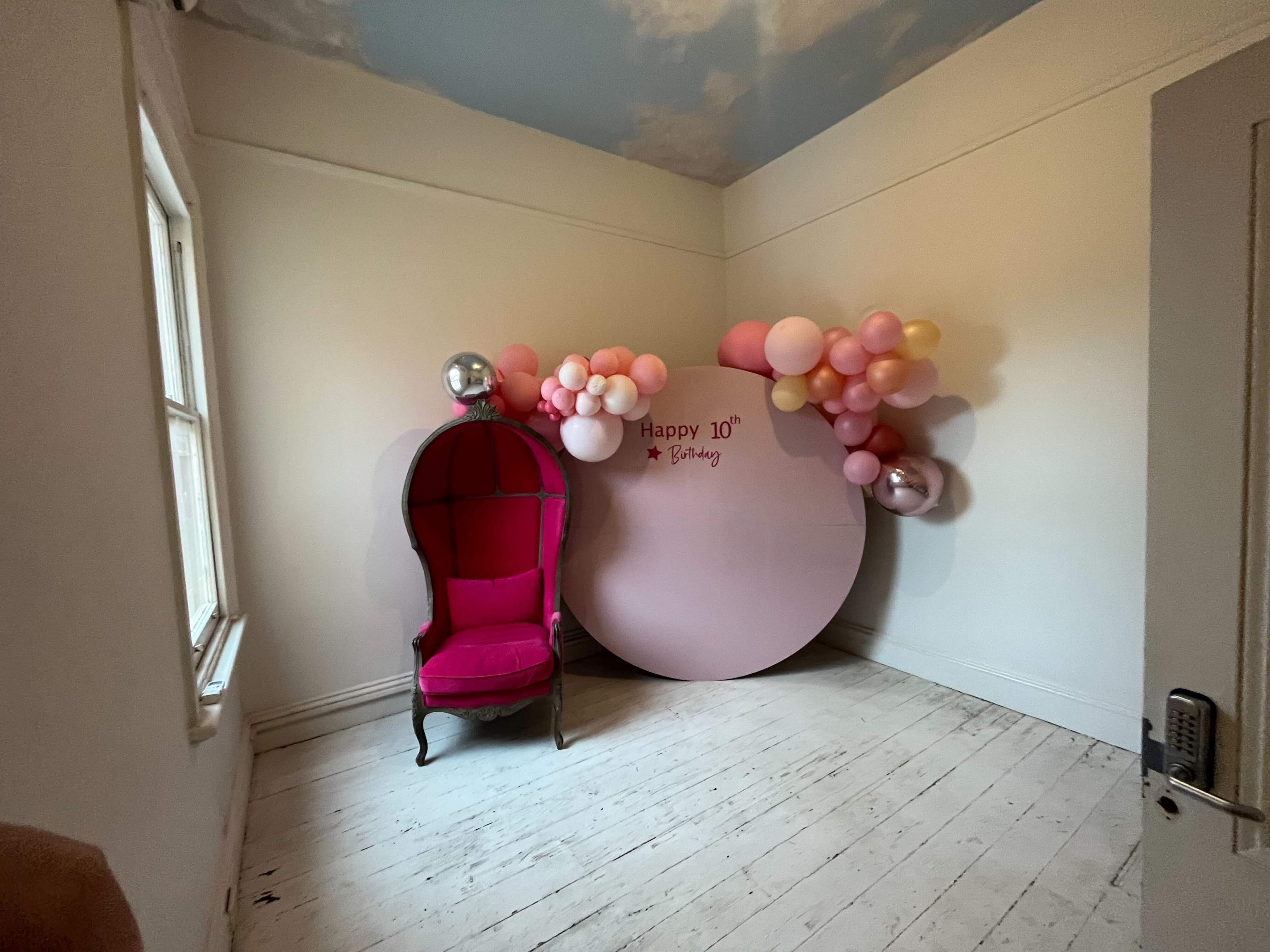 A corner of a room featuring a pink chair and a decorated wall with a round sign that says "Happy 10th Birthday," surrounded by an arrangement of pink and silver balloons.