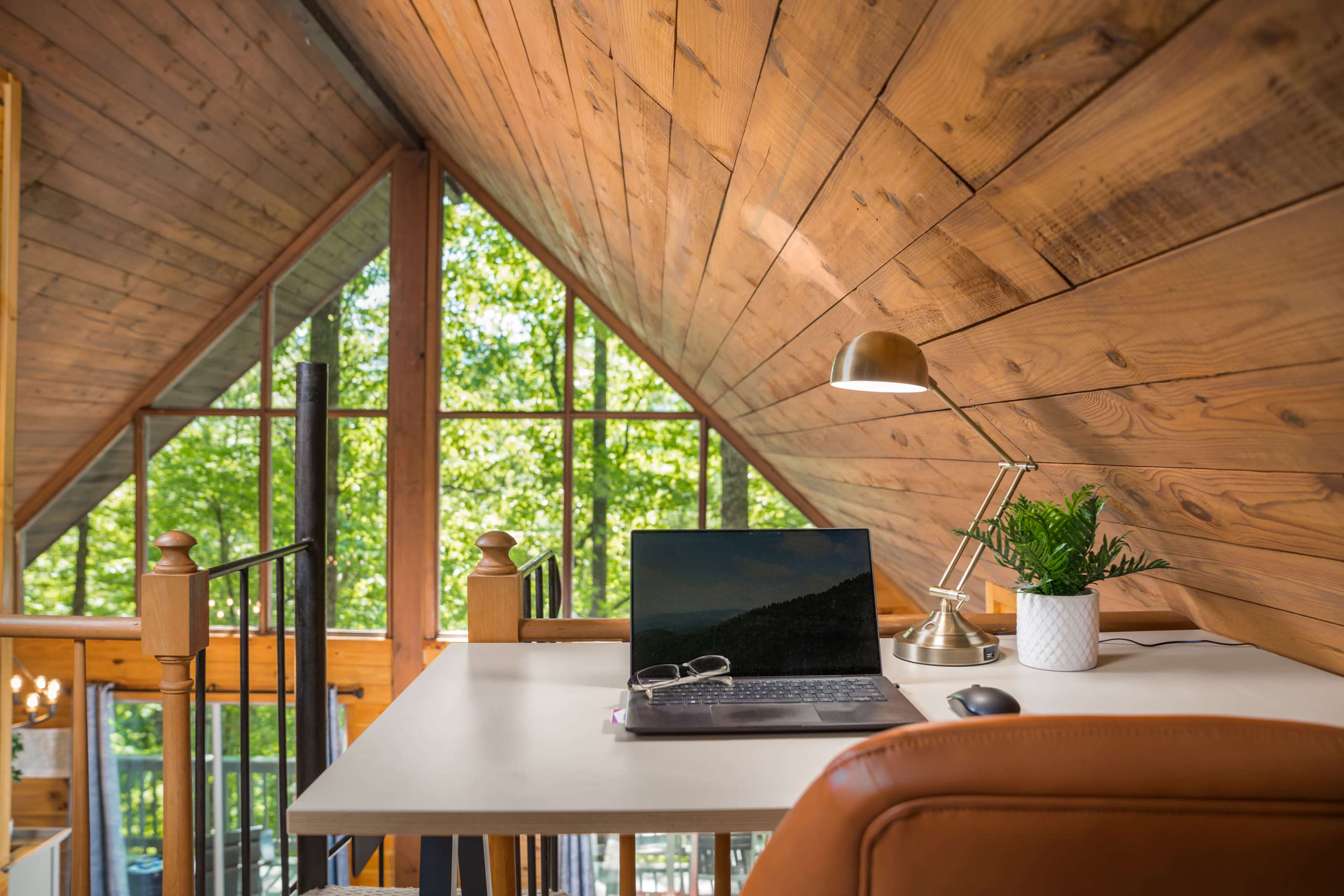 A modern workspace is set up in a loft with wooden walls and large windows letting in natural light, featuring a laptop, a desk lamp, and a small plant.