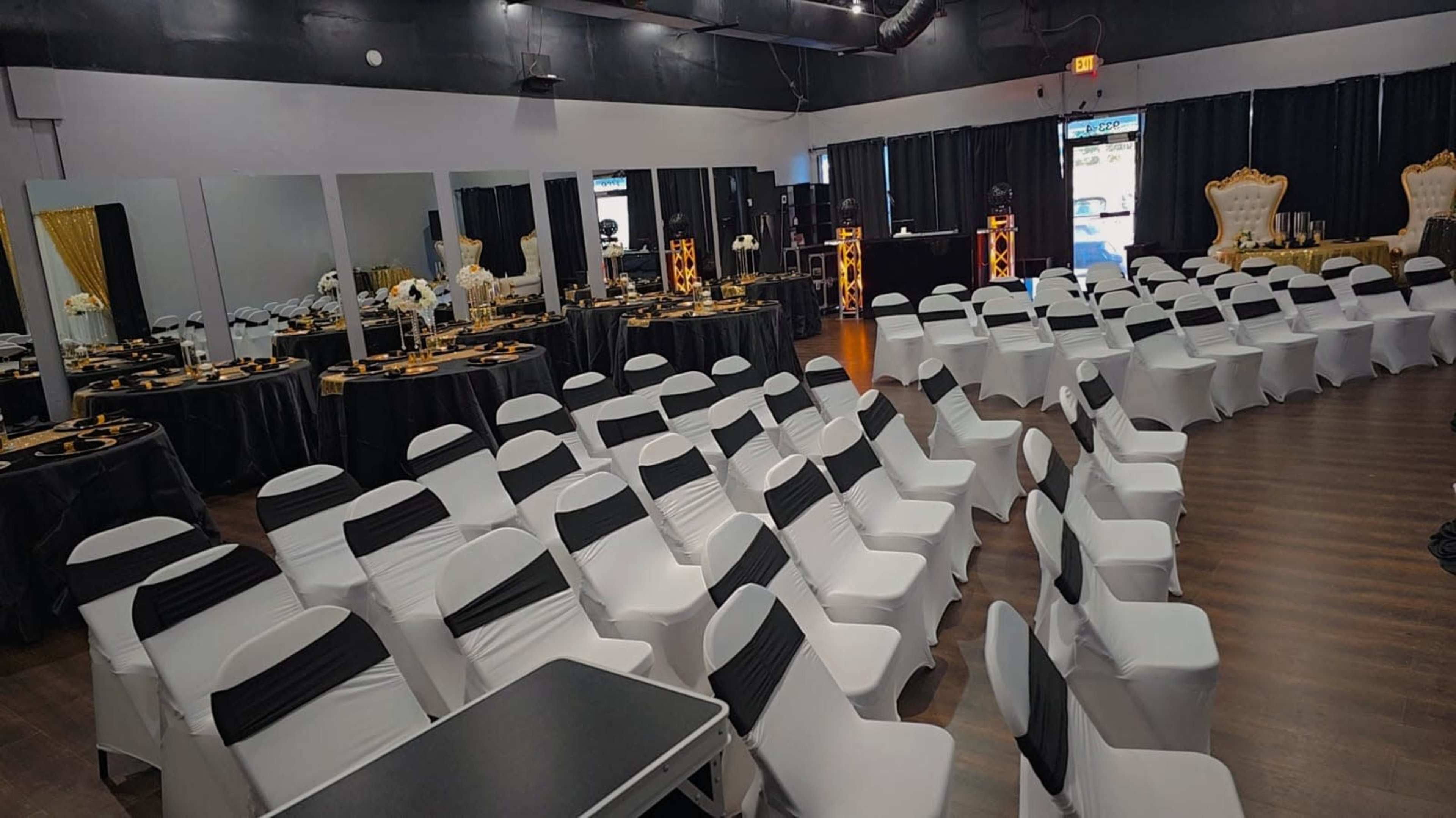 The image shows an event space arranged with white chairs and black tablecloths, set up for a formal gathering with tables decorated along the walls.