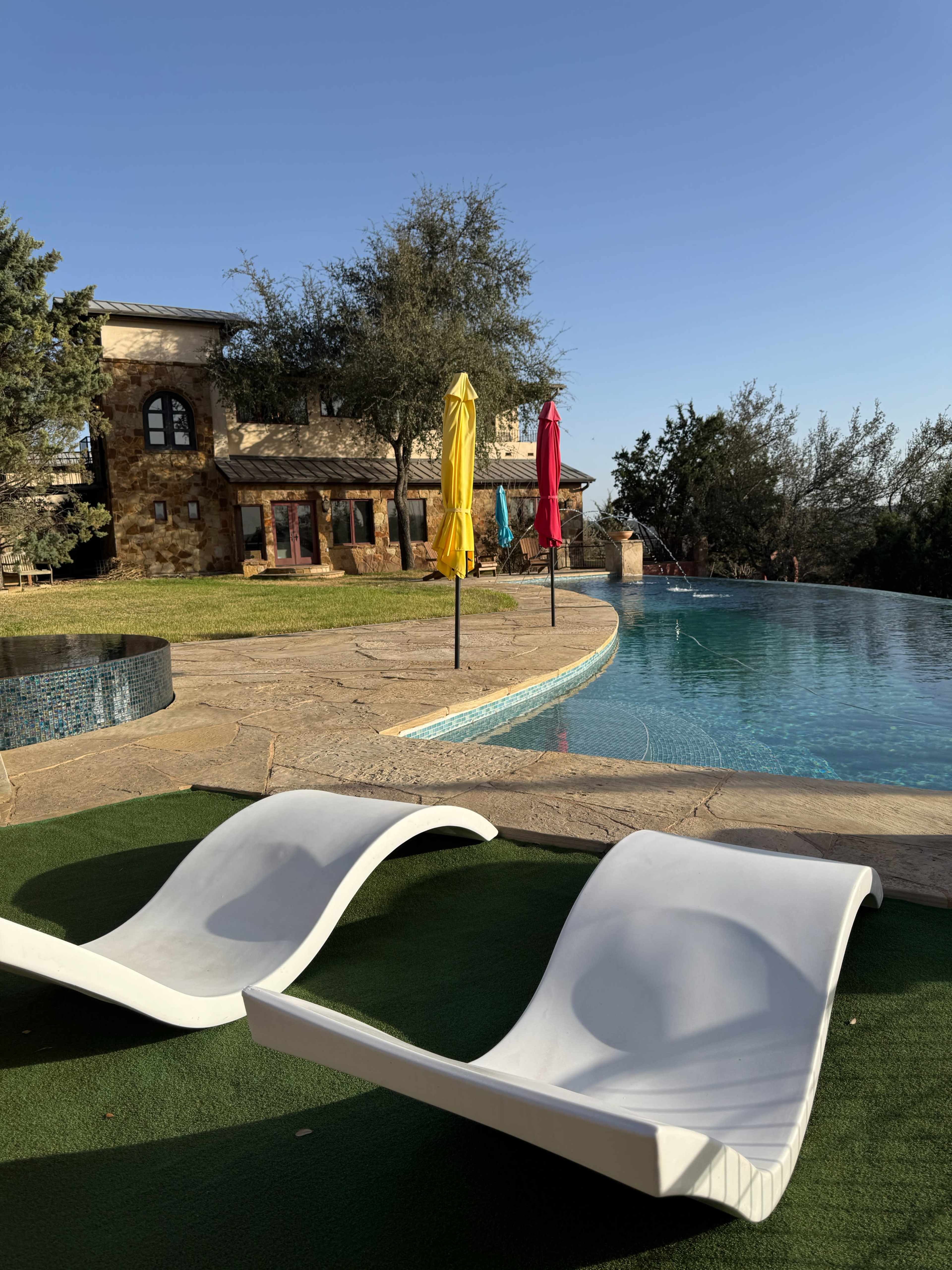 The image shows two white lounge chairs beside a swimming pool with an adjacent stone house and colorful umbrellas in the background.