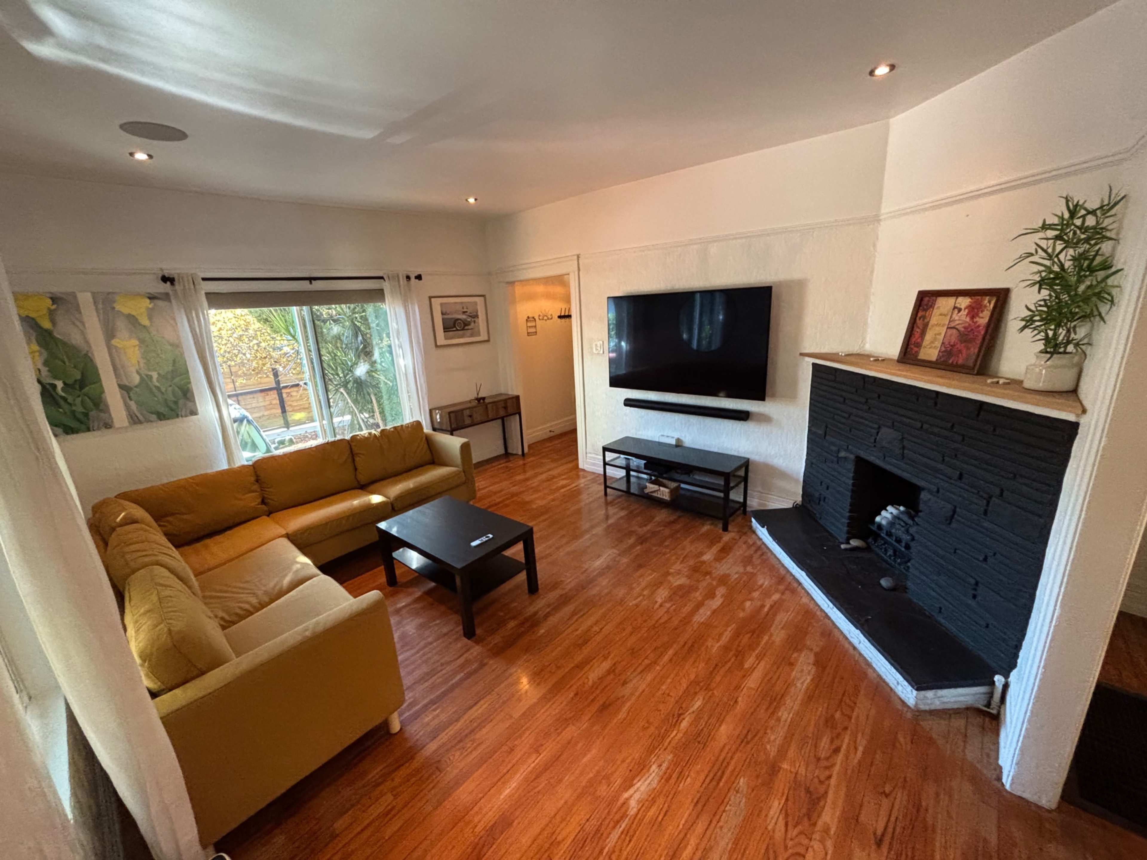 The room features a yellow couch, a black fireplace, a flat-screen TV mounted on the wall, and a coffee table, with large windows letting in natural light.