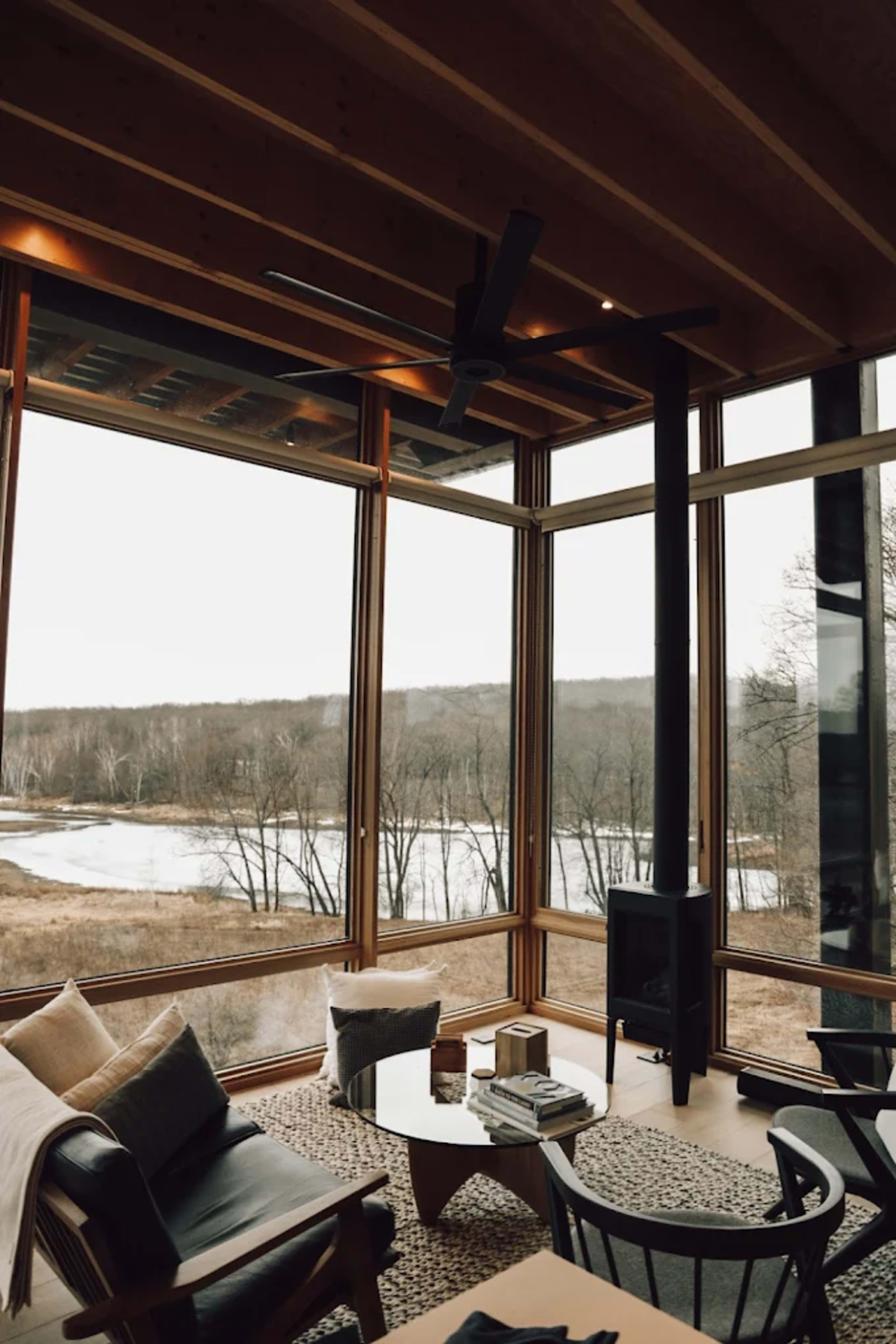 The image shows a modern living space with large windows overlooking a river and trees, featuring a cozy seating arrangement and a wood stove.