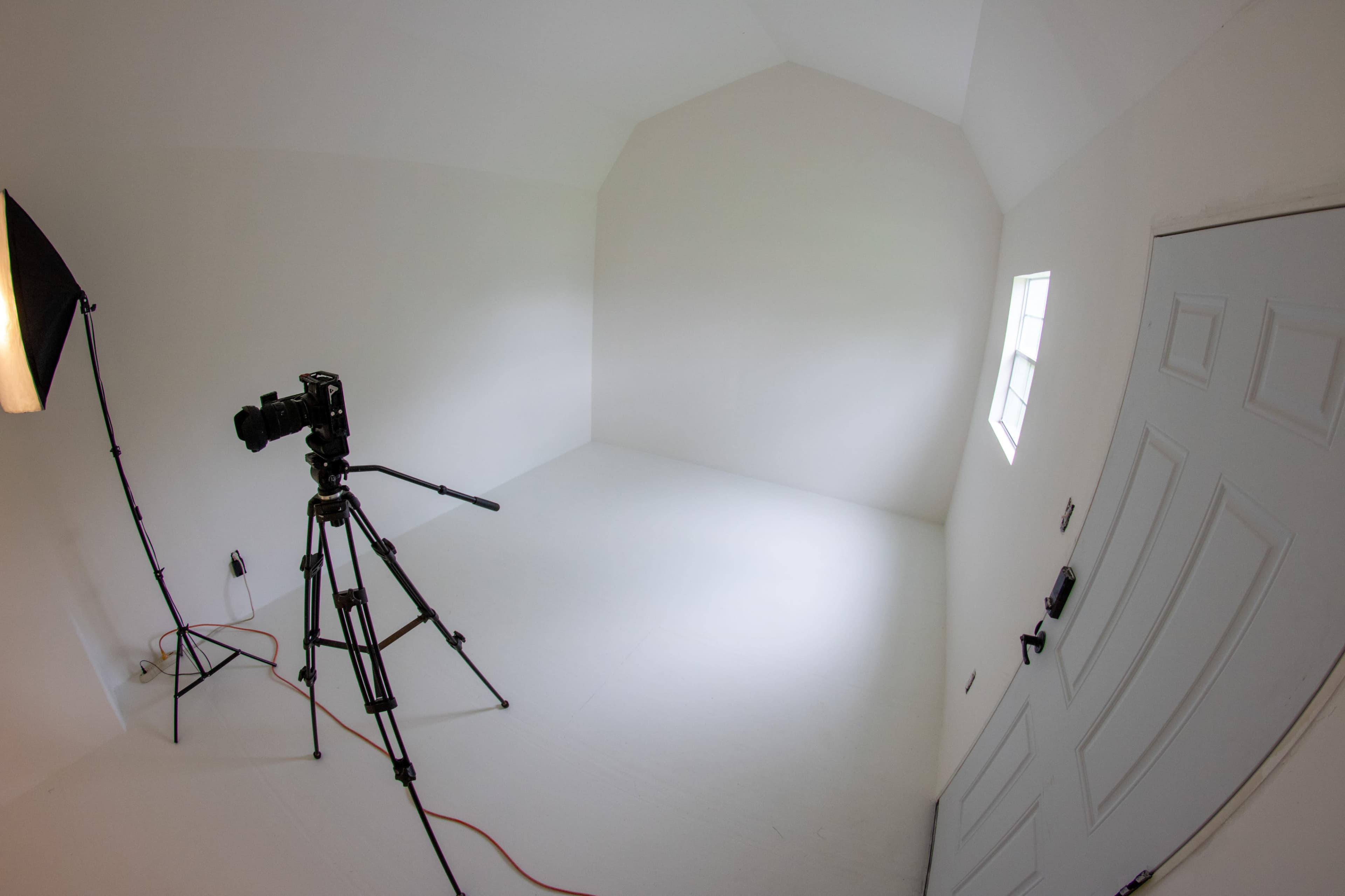 The image shows an empty, well-lit room with white walls and a camera set on a tripod facing a door.