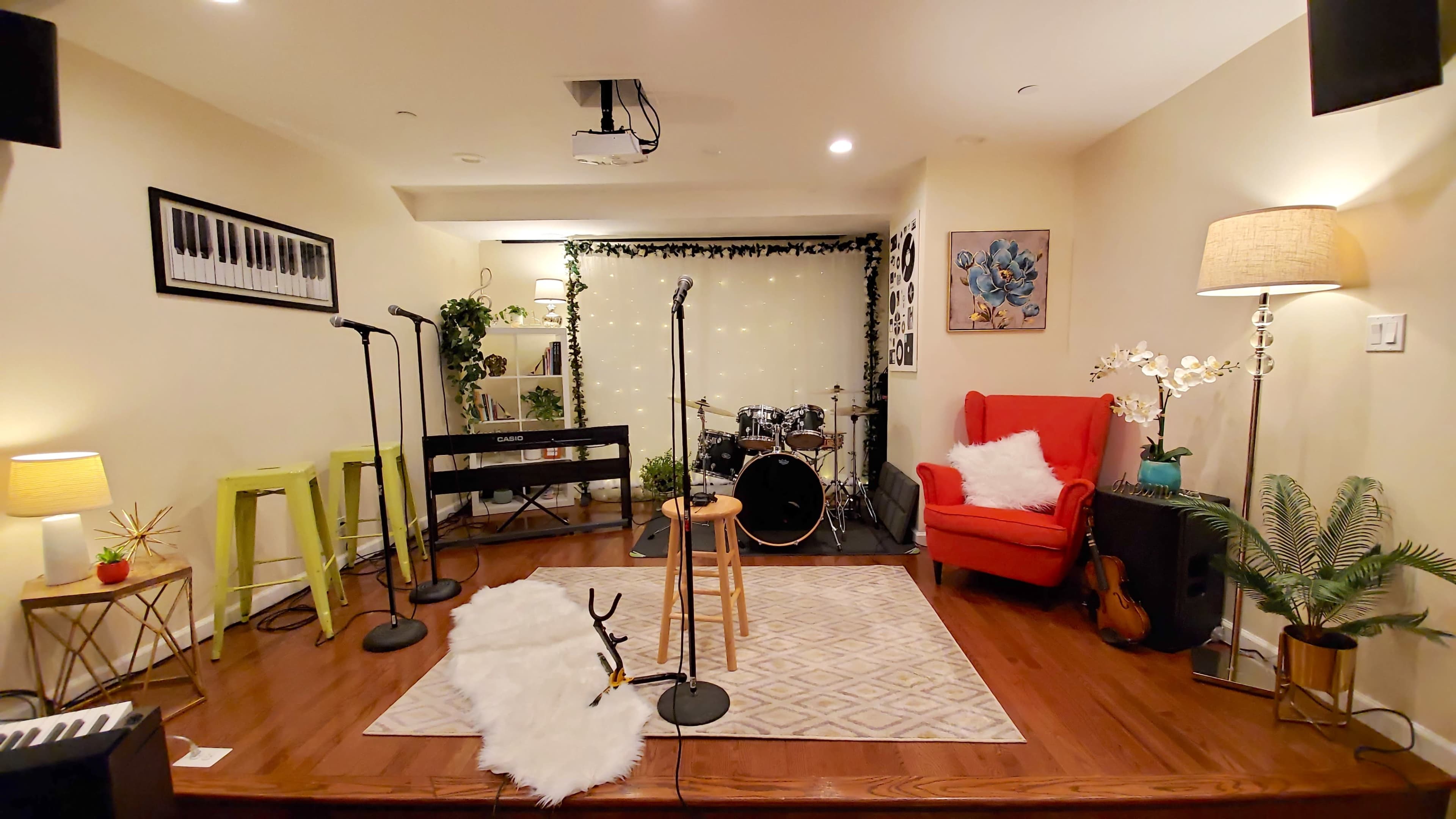 A small performance space featuring microphones on stands, a drum set, a piano, and seating arranged around a rug.