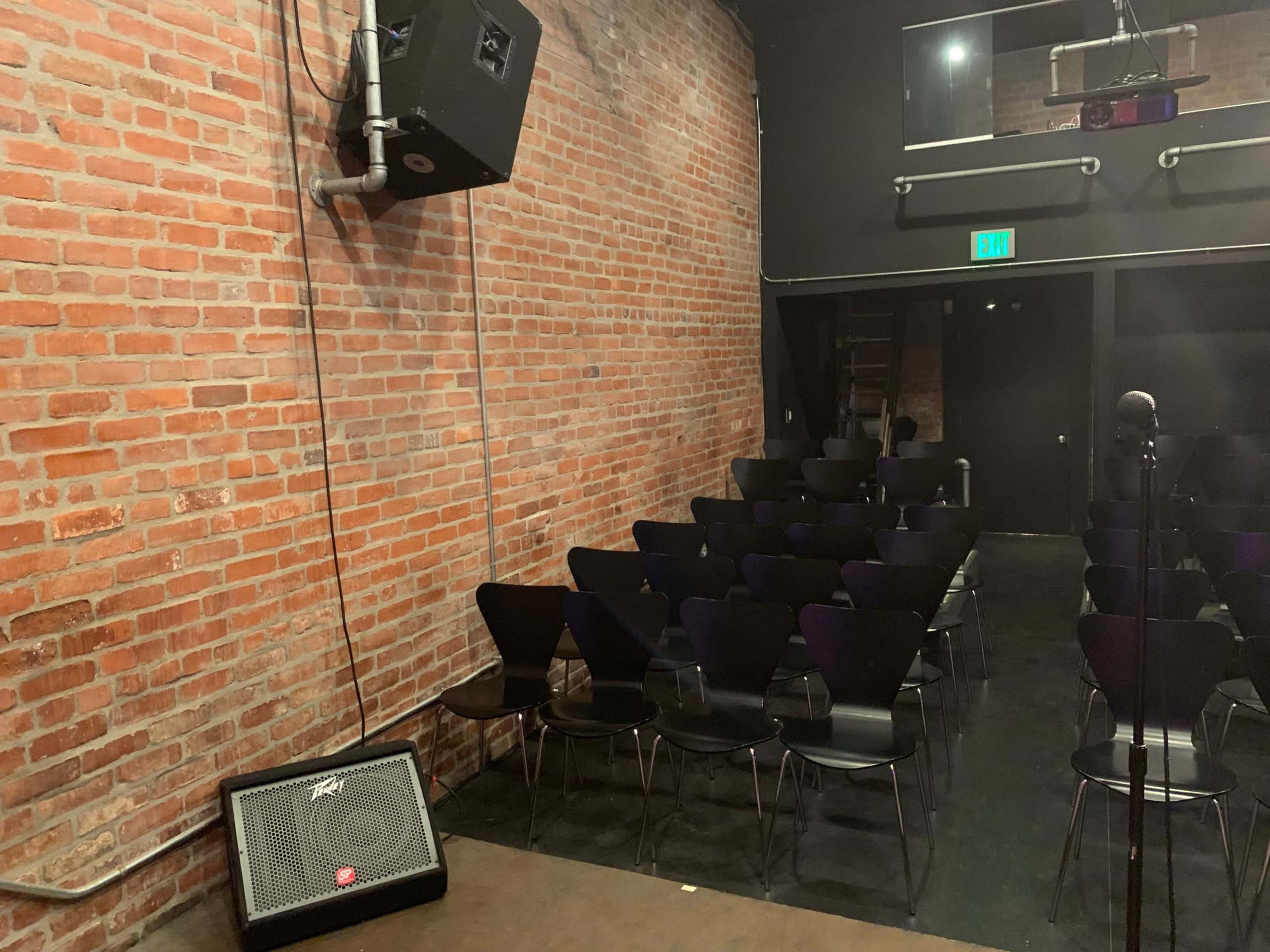 An empty performance space with a brick wall, a microphone stand, and rows of black chairs facing a small stage area.