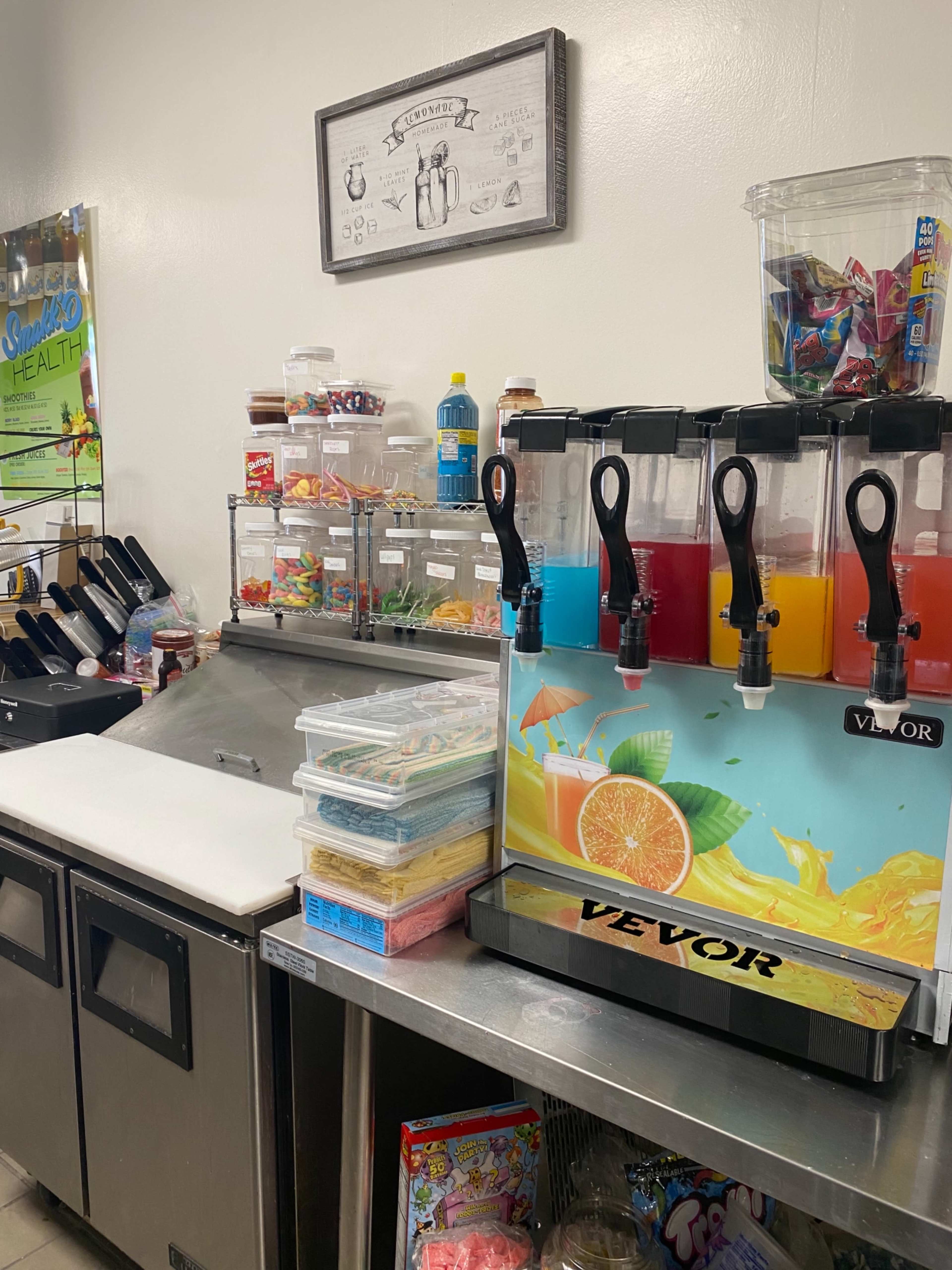 A beverage dispenser with multiple colorful drink options next to a countertop filled with various snacks and condiments.