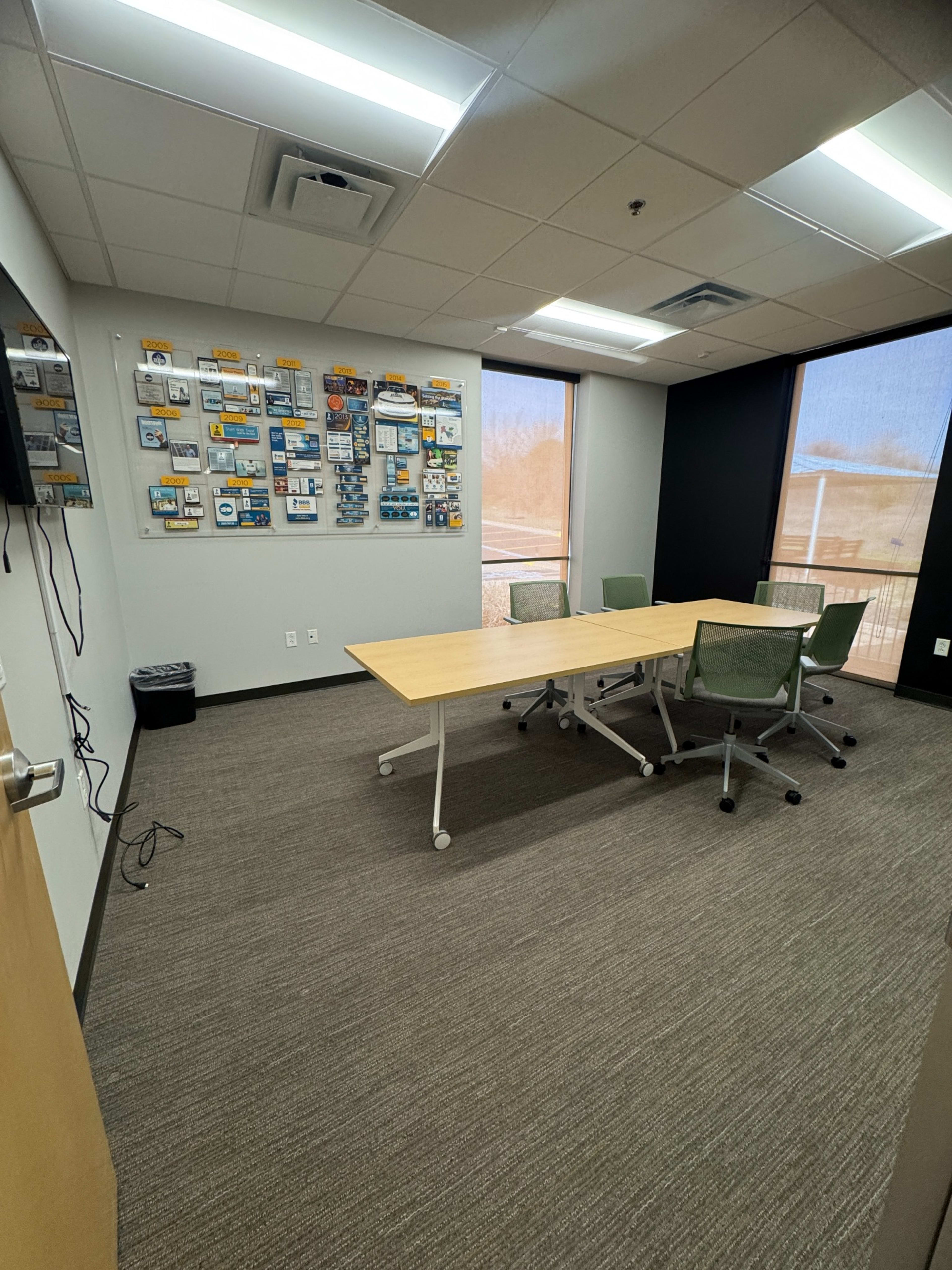 The image shows a small meeting room with a rectangular table, four chairs, and a wall-mounted display of various items, along with a window allowing natural light.