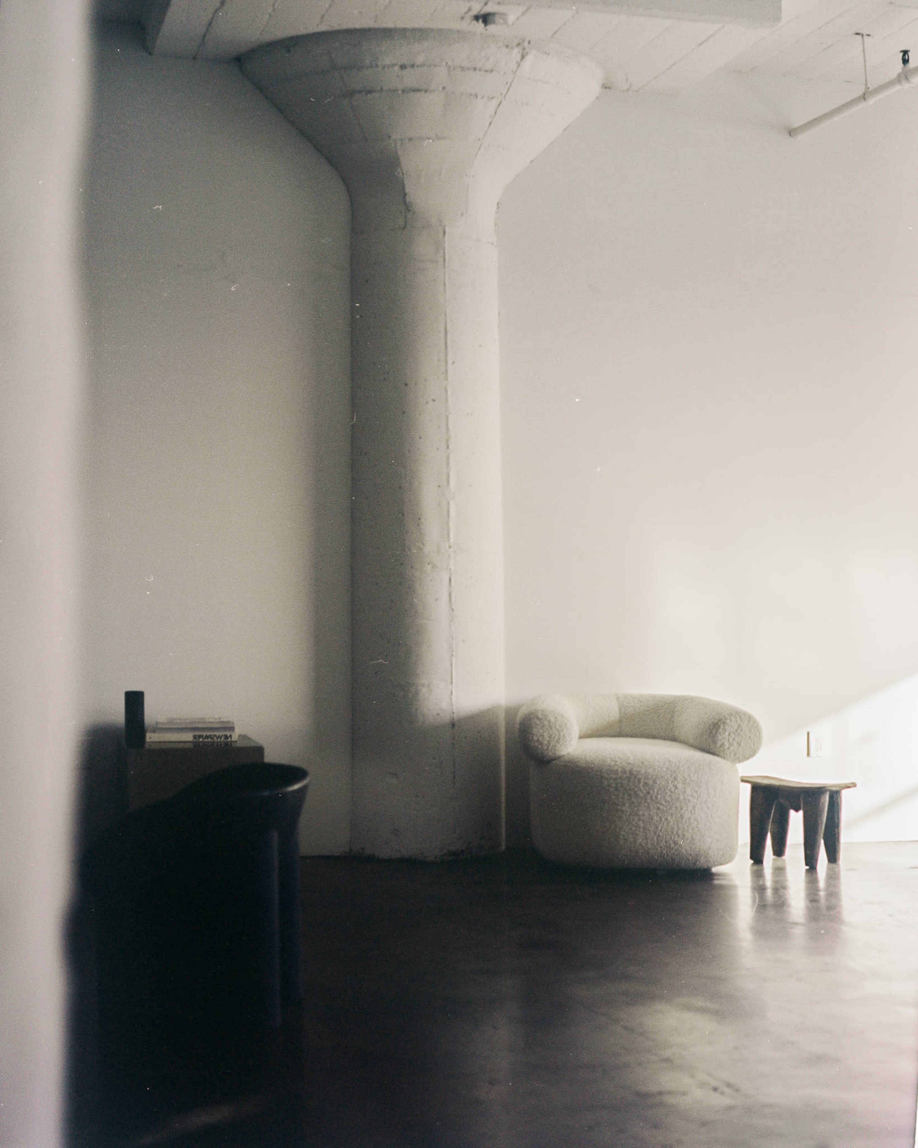 The image shows a minimalist interior with a textured white chair, a small wooden table, and a large concrete pillar against a white wall.