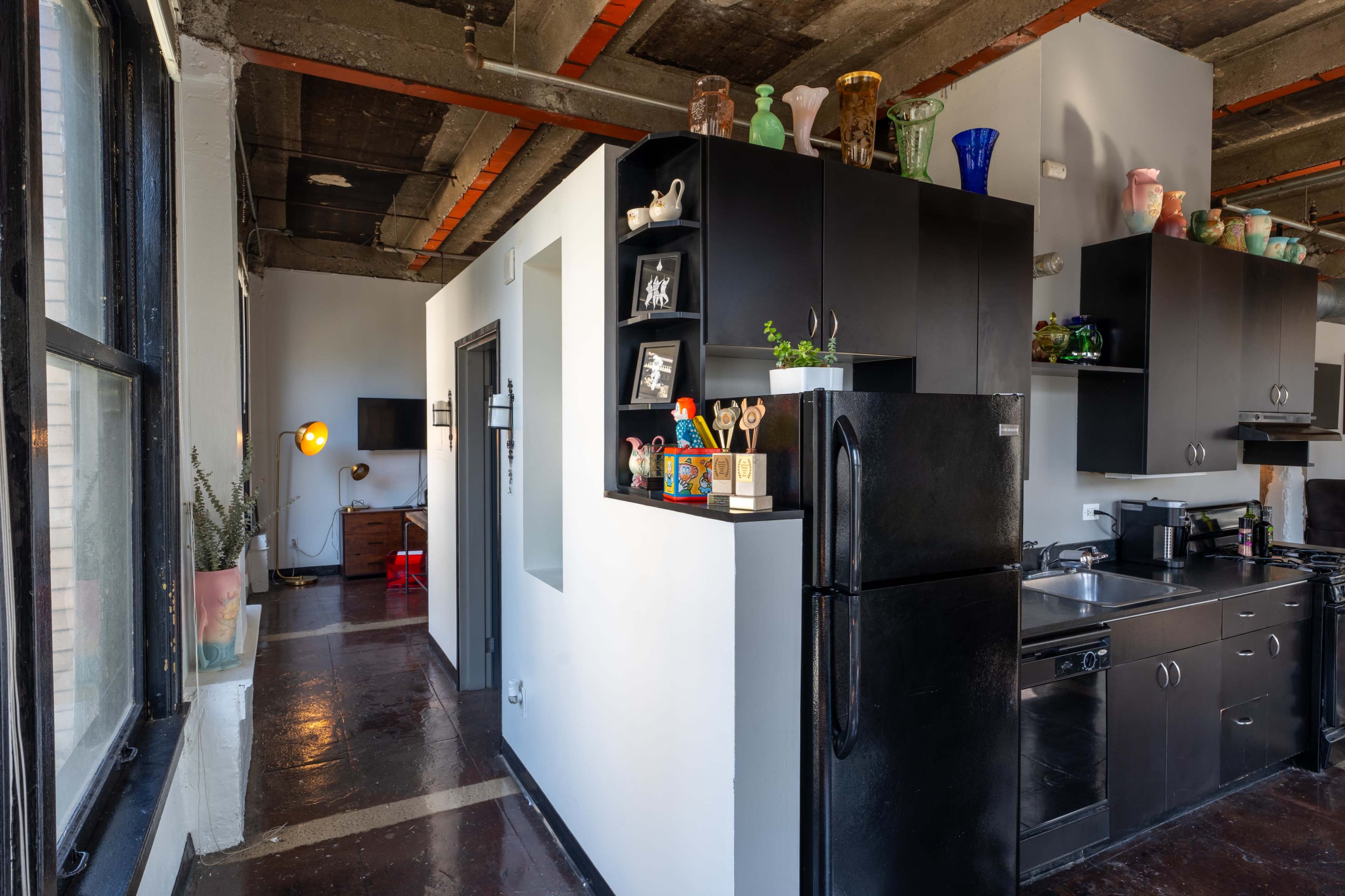 A modern kitchen with black cabinets and appliances, adjacent to a small hallway leading to a living area.