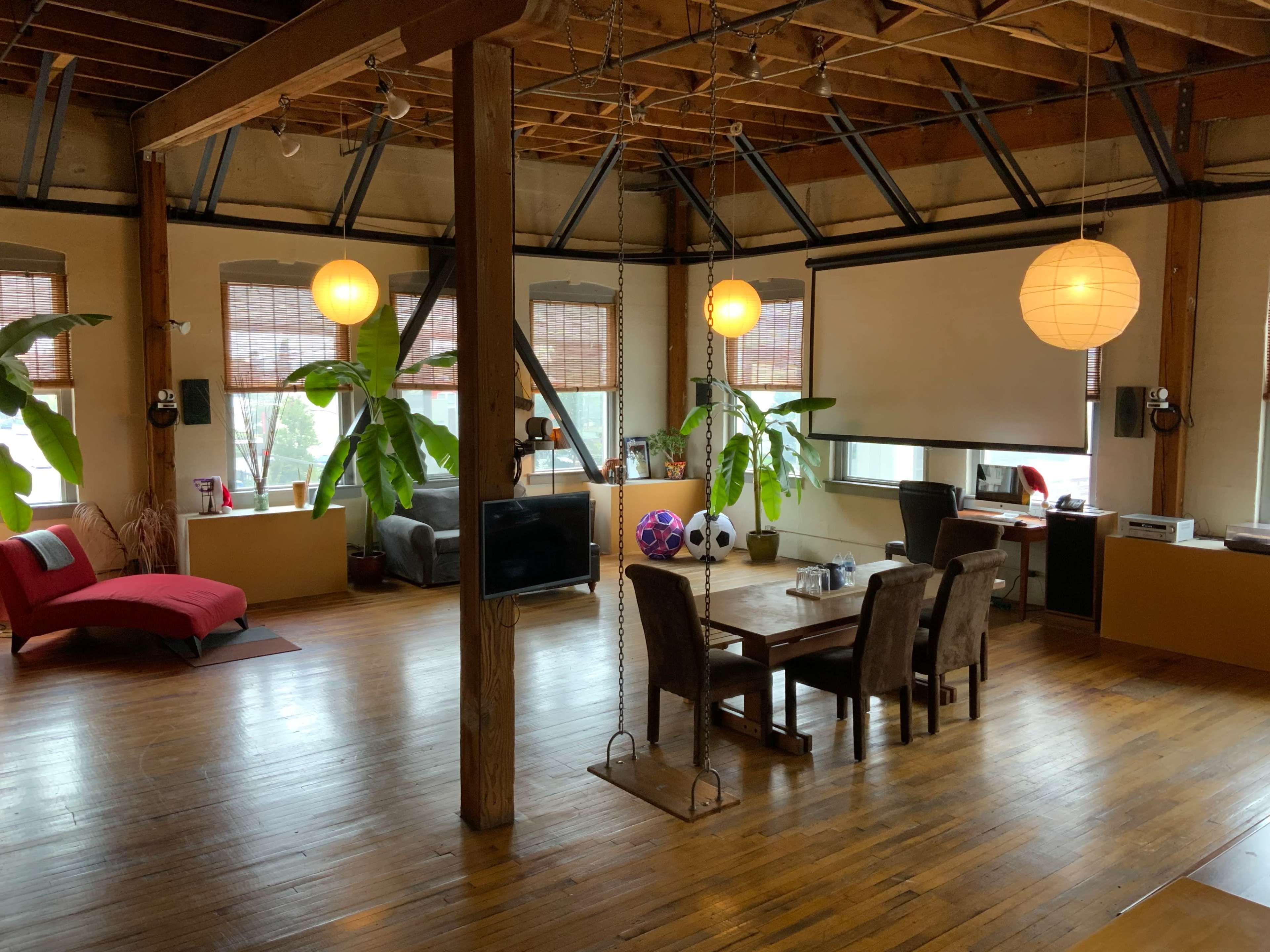 A spacious loft interior featuring wooden floors, large windows, a dining table, and seating areas with plants and decorative lighting.
