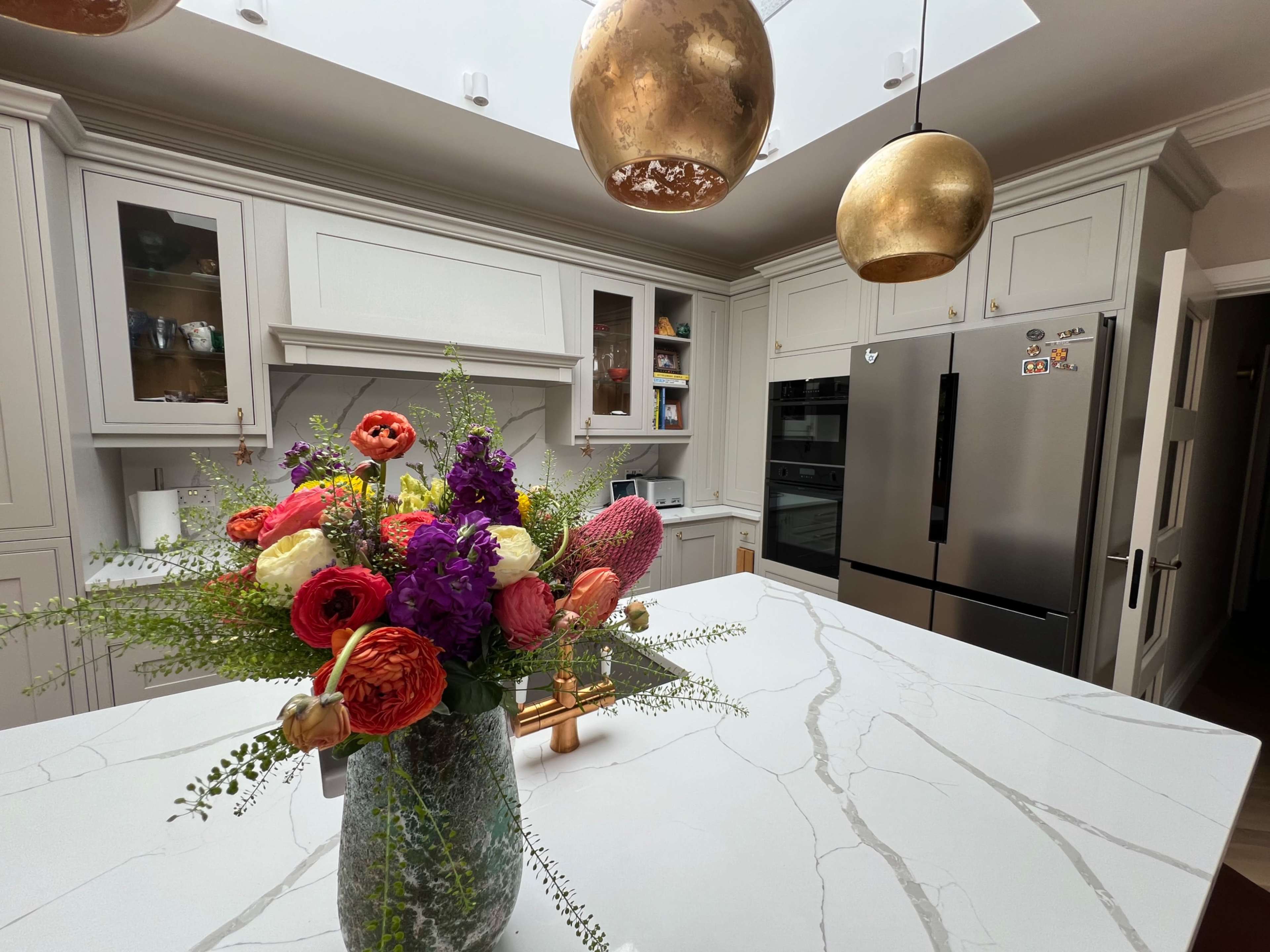 A modern kitchen with a light-colored marble countertop, a floral arrangement in a vase, and two golden pendant lights hanging above.