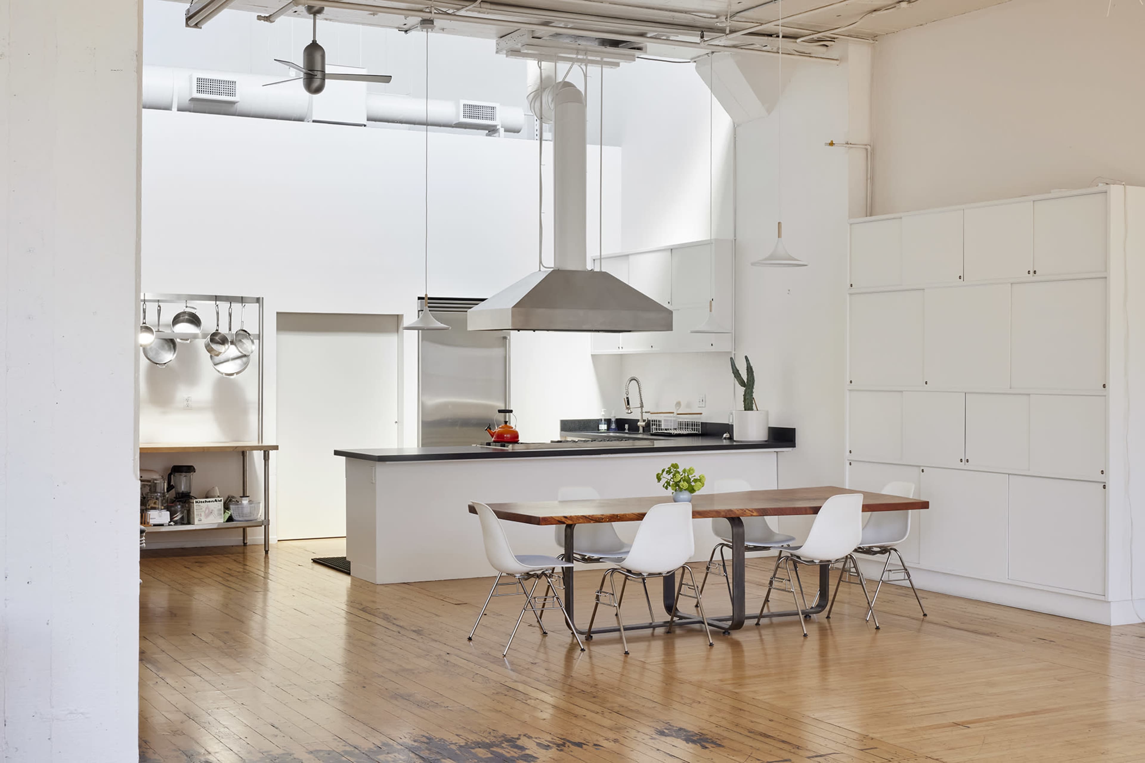 A modern kitchen features a large wooden dining table surrounded by white chairs, with stainless steel appliances and a prominent range hood above the cooking area.
