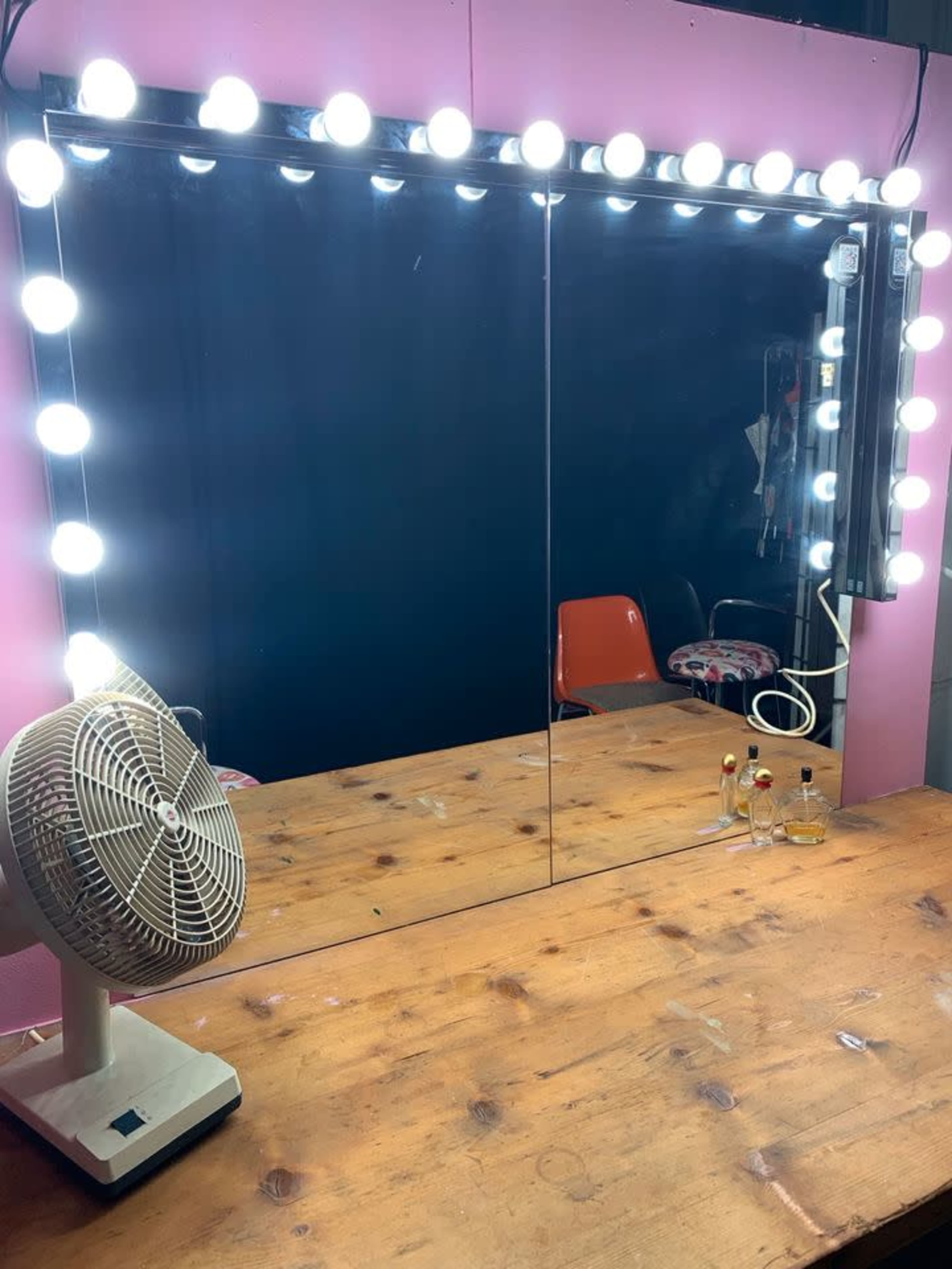 A well-lit vanity mirror surrounded by light bulbs, with a fan and a few bottles placed on a wooden table.