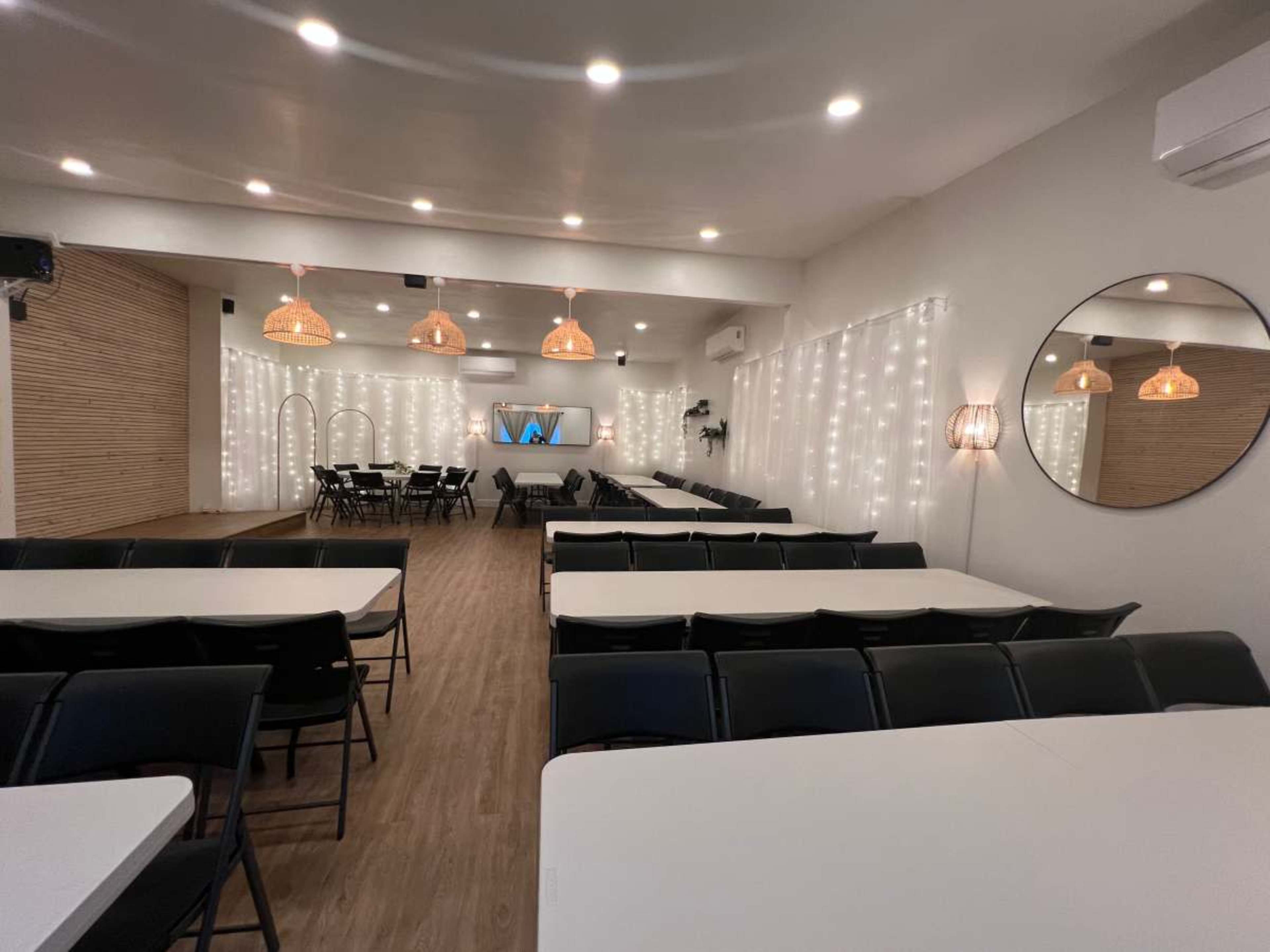 A spacious room with rectangular tables and black chairs arranged for a gathering, accented by string lights and decorative light fixtures.
