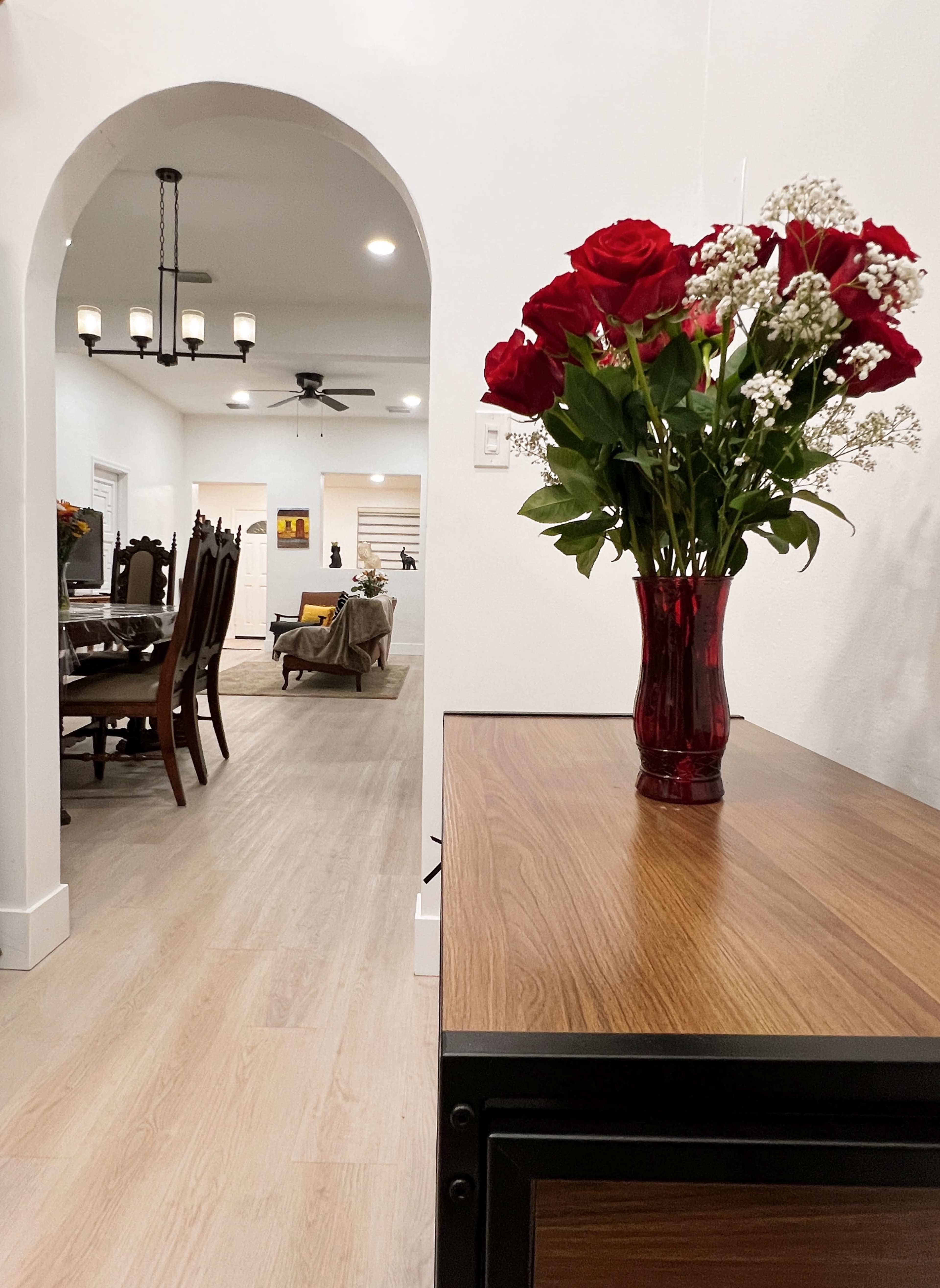 A vase of red roses and white fillers sits on a wooden table, with an arched entryway leading into a dining area and living space.