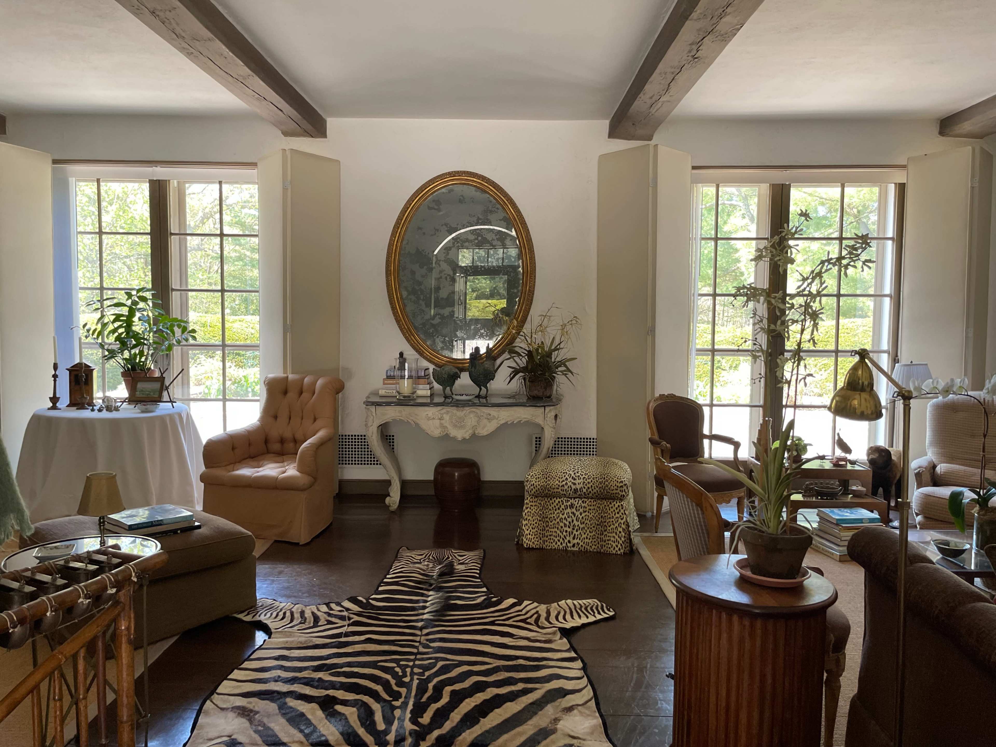 The image shows a bright living room with large windows, featuring a mix of vintage furniture, a decorative mirror, a zebra-striped rug, and several plants.