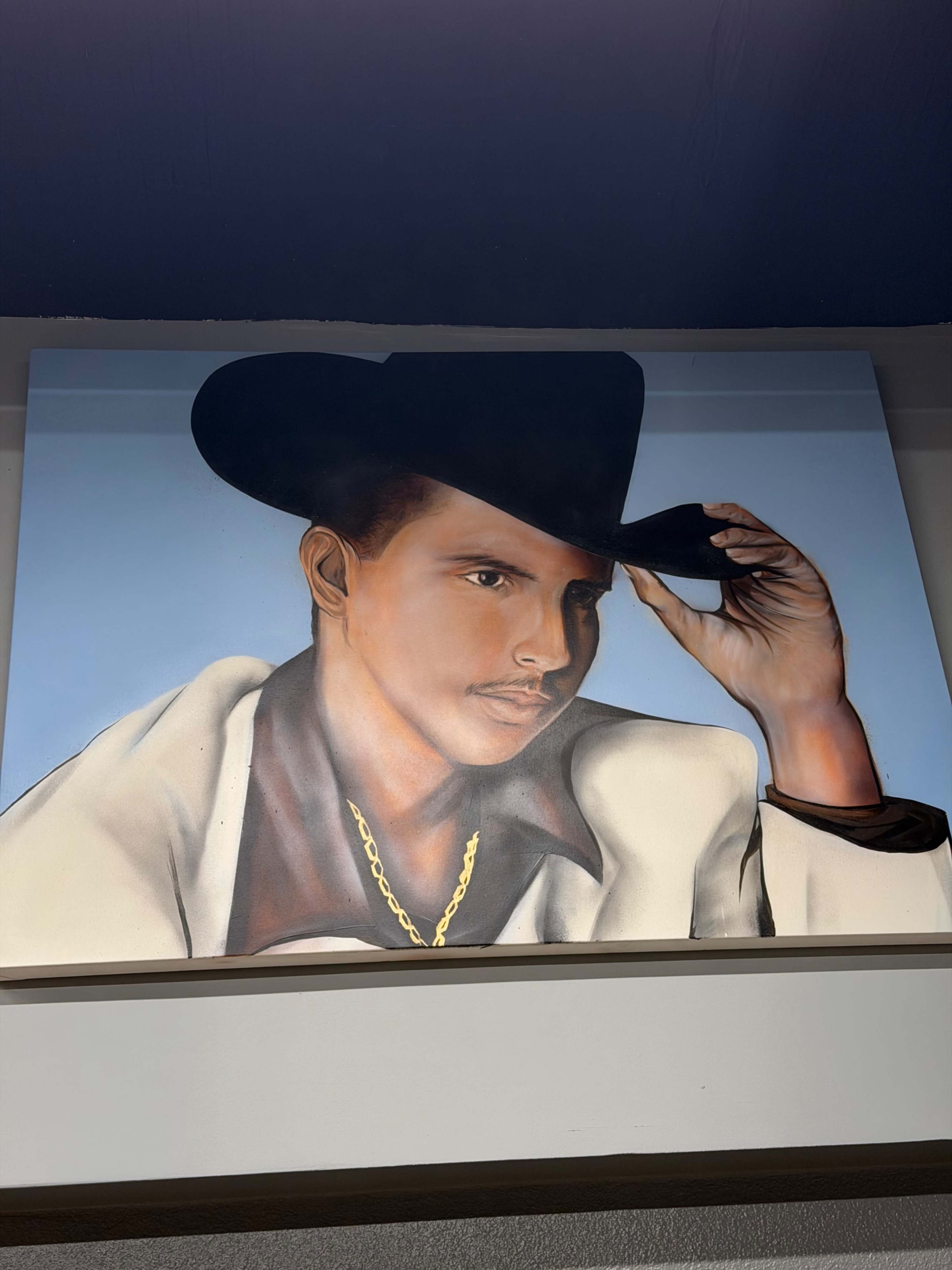 The image depicts a painted portrait of a young man wearing a cowboy hat and a light-colored suit, with his hand holding the brim of the hat.