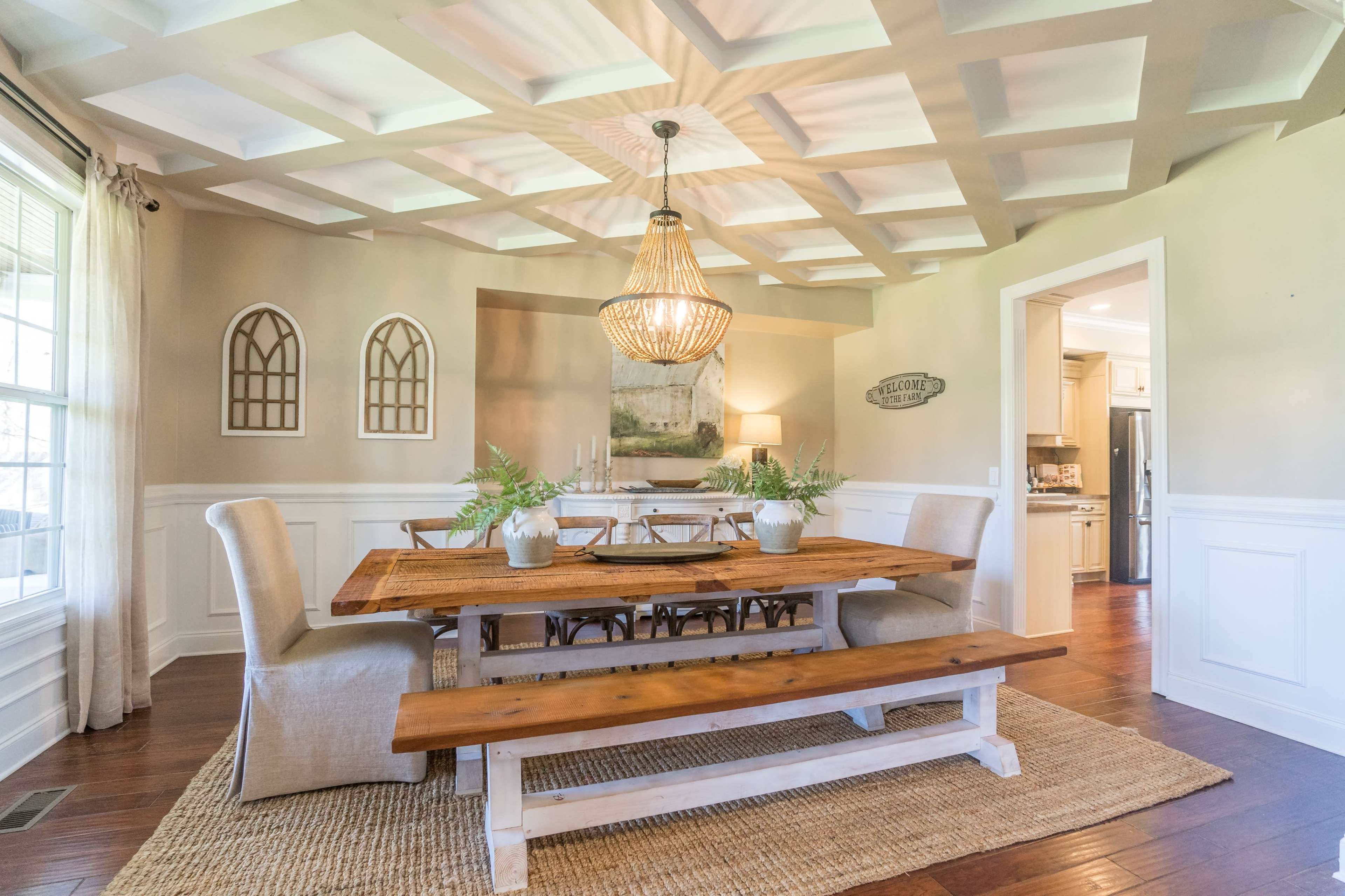 A spacious dining room features a wooden table with benches, a chandelier above, and decorative wall accents, with a doorway leading to a kitchen.