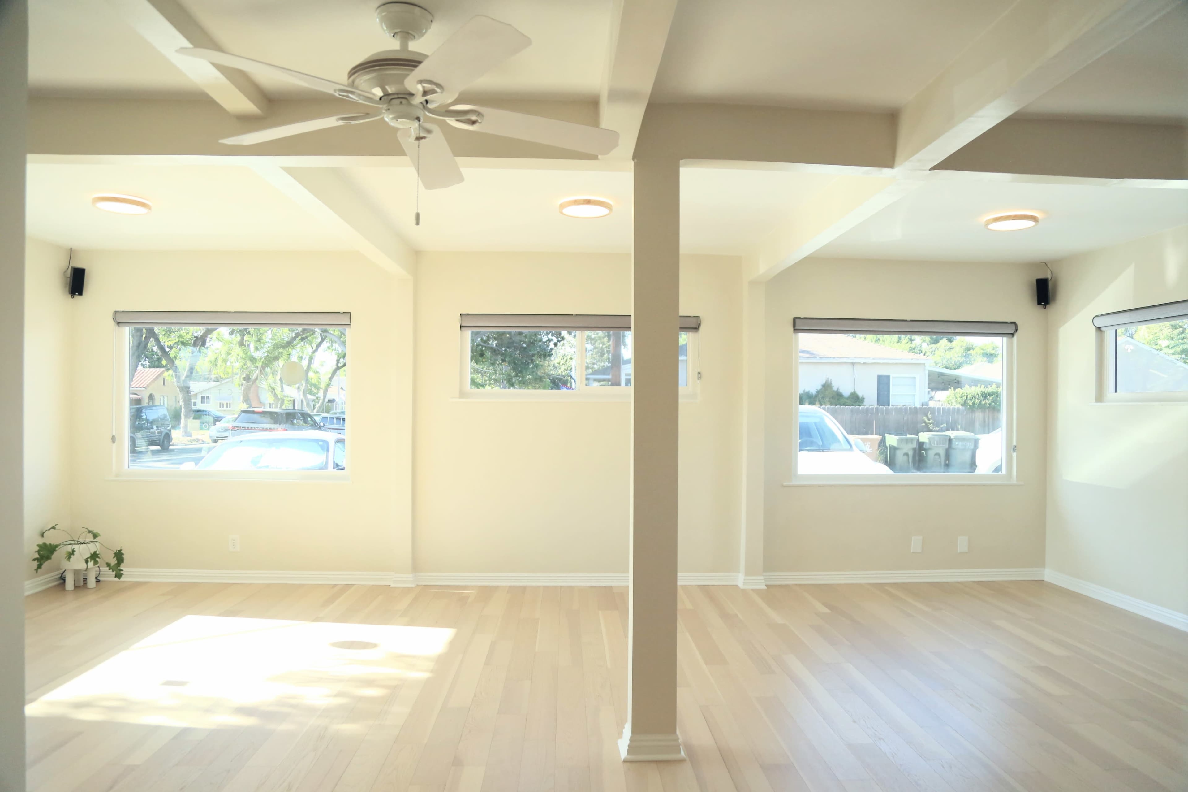 The image shows a spacious, well-lit room with light wood flooring, large windows, a ceiling fan, and a central support column.