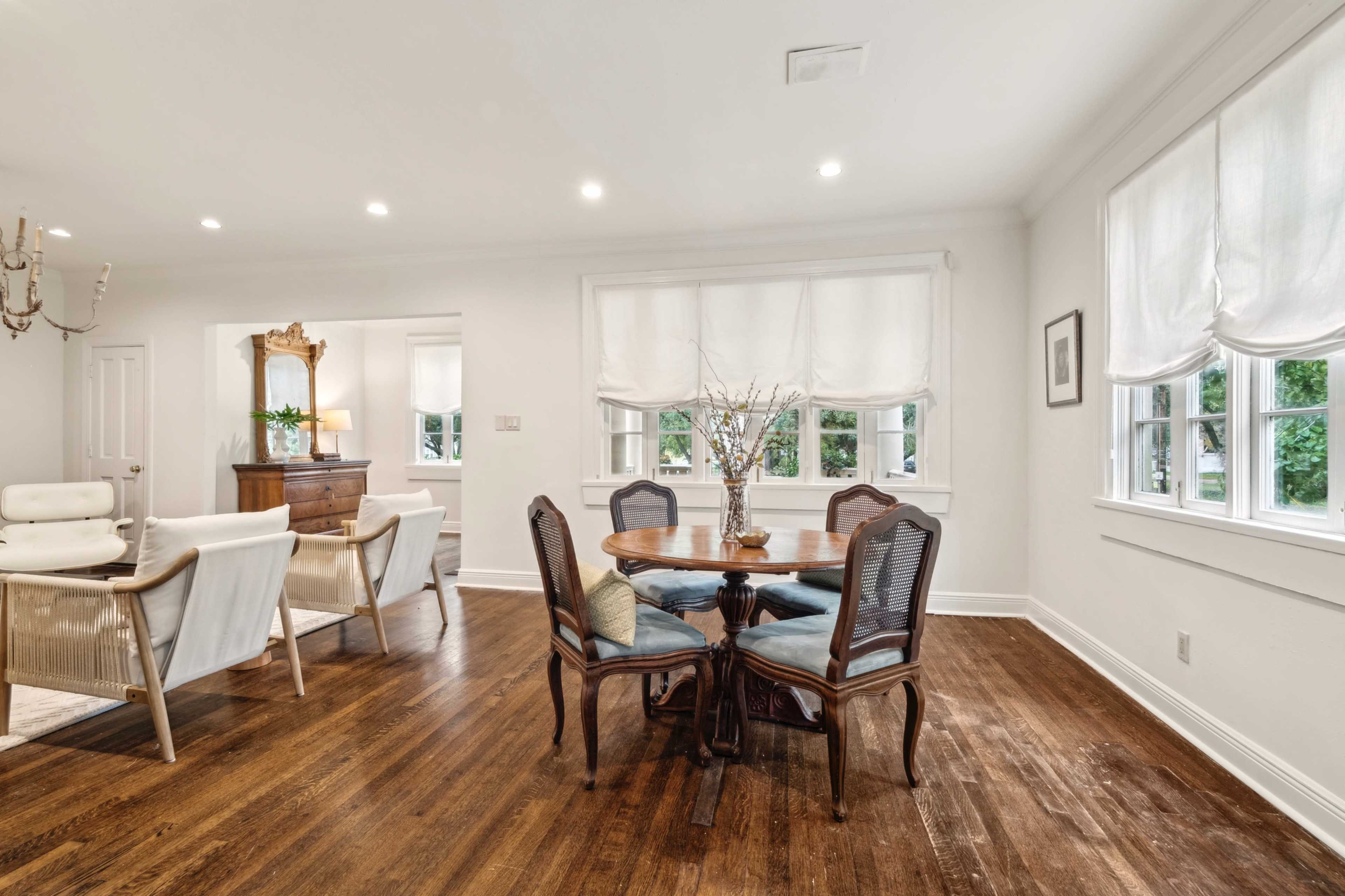 A dining area features a round wooden table with four chairs, surrounded by large windows with light-filtering shades, and is adjacent to a well-lit living space.
