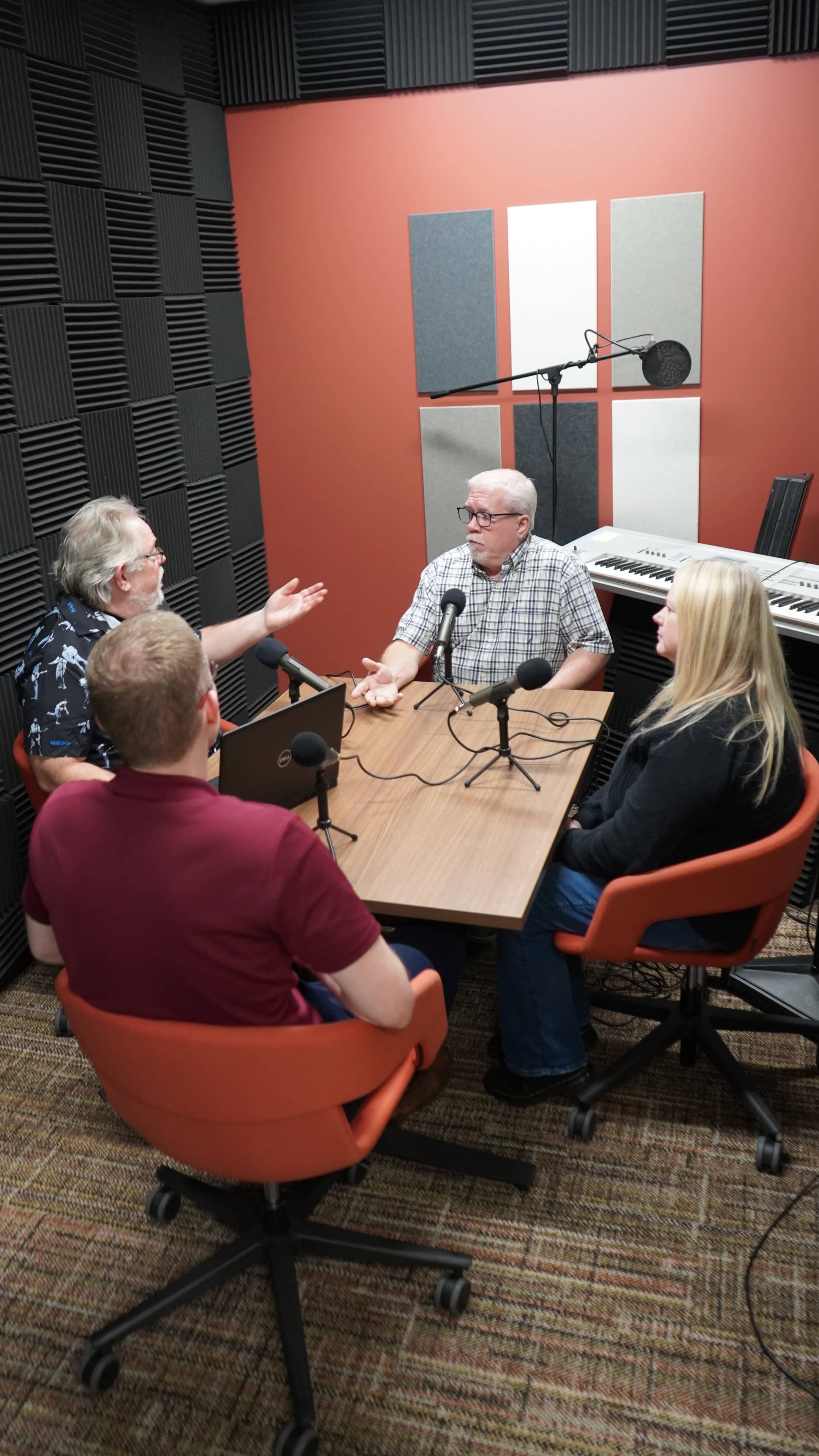 A group of four people engaged in discussion around a table in a podcast studio, with microphones positioned in front of them.