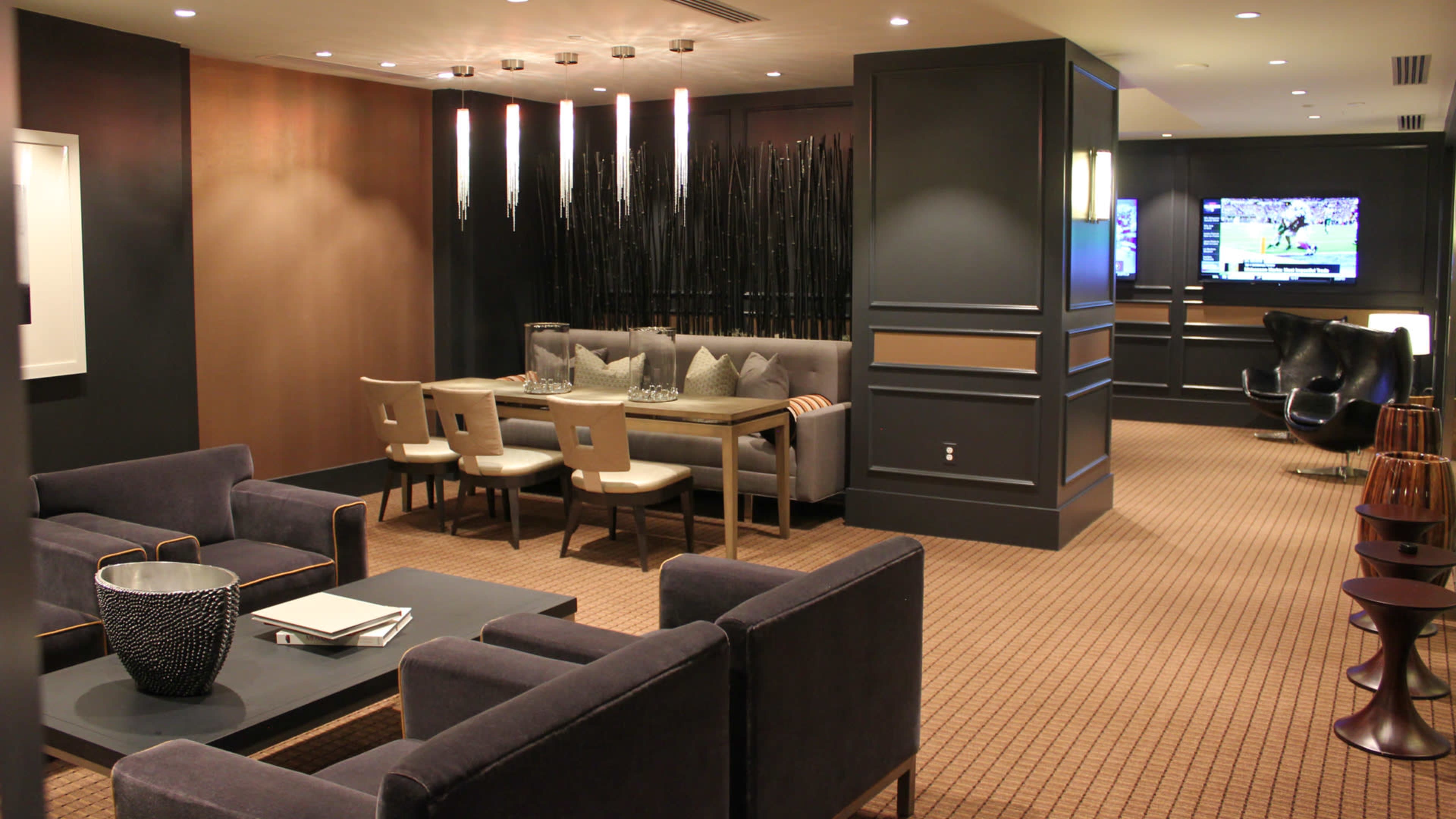 The image shows a modern lounge area featuring seating arrangements, a dining table, decorative lighting, and a television mounted on the wall.