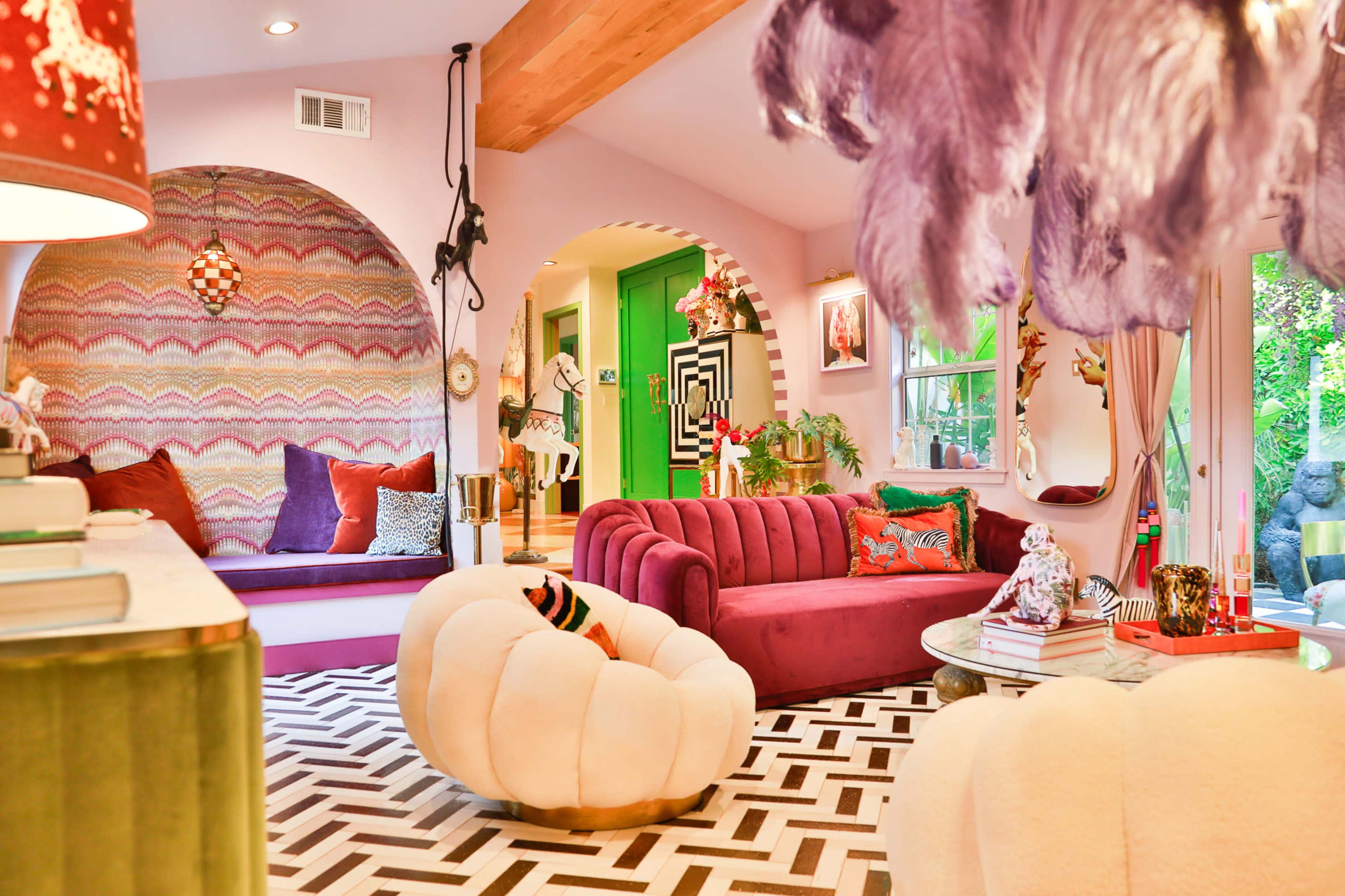 The image shows a colorful living room featuring a pink and purple color scheme, plush seating, geometric patterns on the floor, and vibrant decor elements.