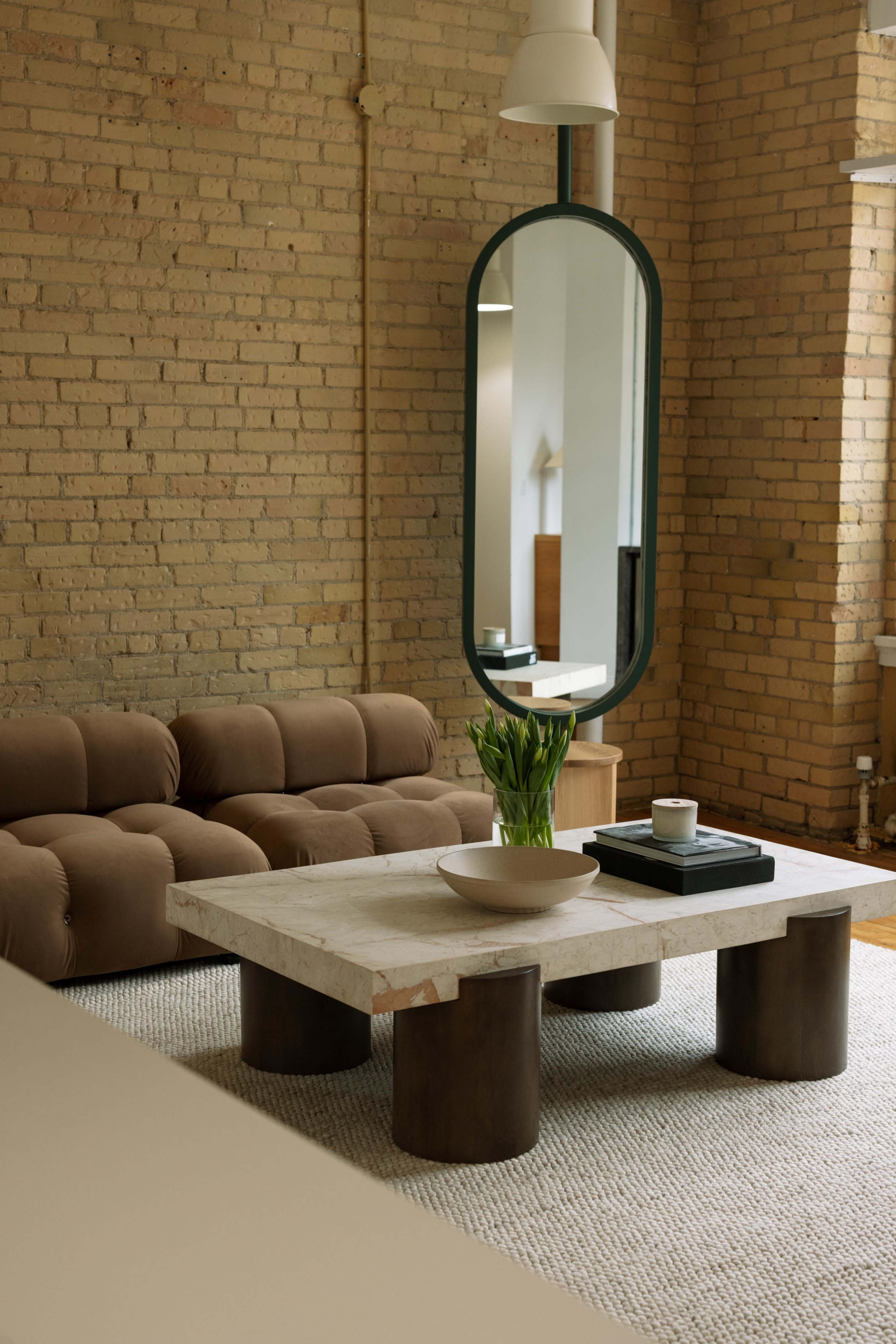 The image depicts a cozy living room featuring a brown tufted sofa, a round mirror hanging above a stone coffee table, and a vase with fresh flowers.