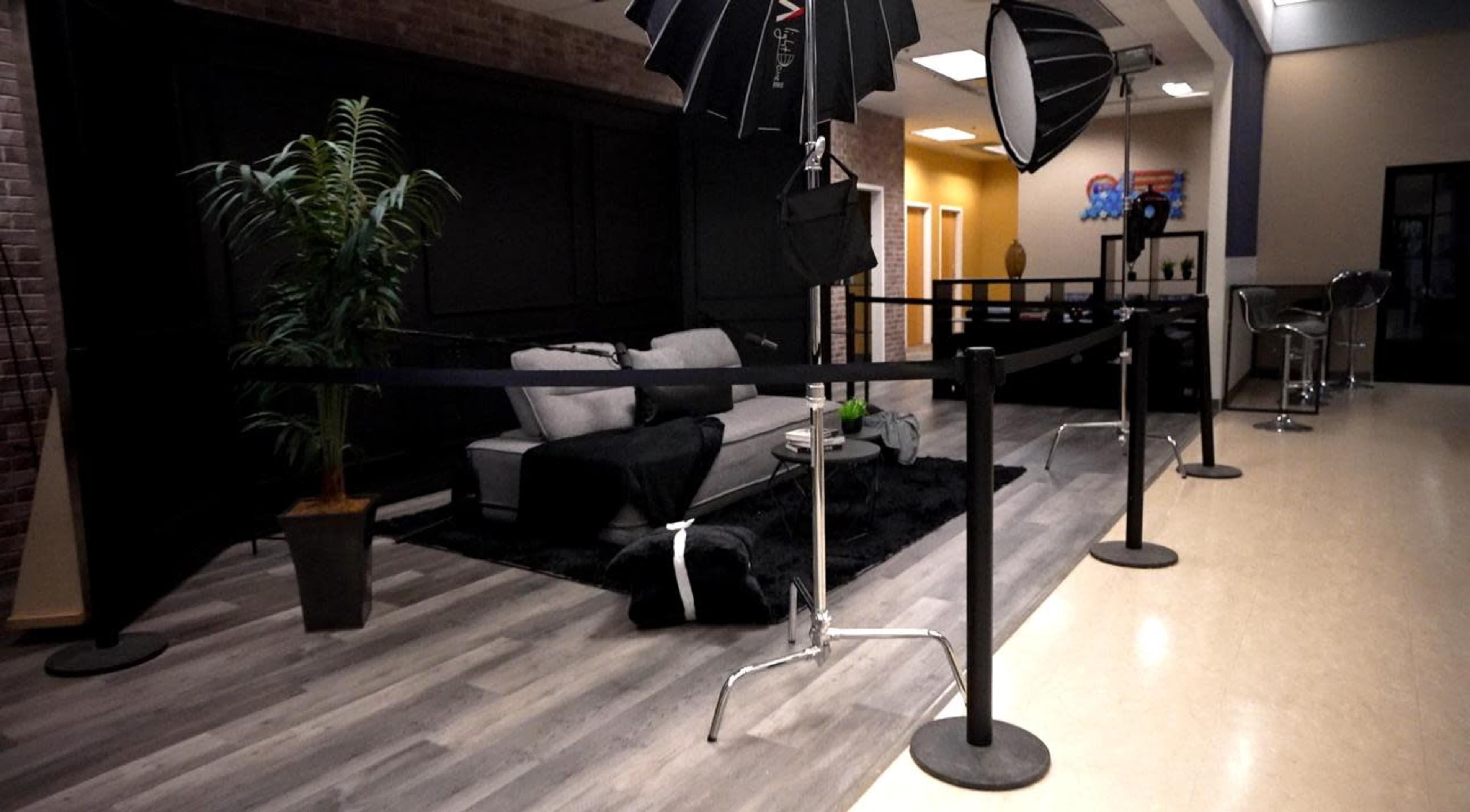 The image shows a photography setup featuring a gray sofa, studio lights on stands, a potted plant, and a black rug in a spacious indoor area with a reception-like atmosphere.